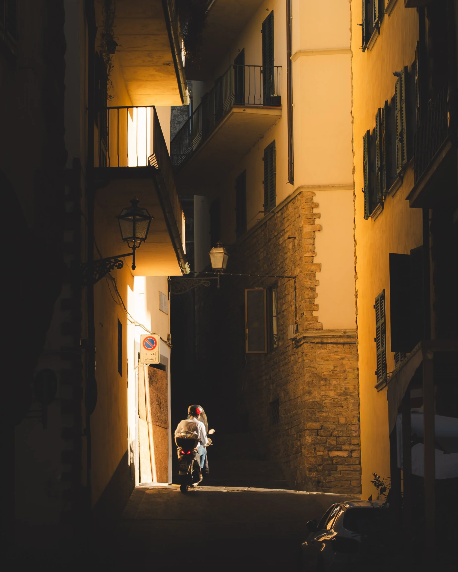 A narrow street in an Italian town with yellow and beige buildings, a person riding a scooter up a steep incline, and parked cars on the side. Lionel Chu - Hotel & Travel Photographer