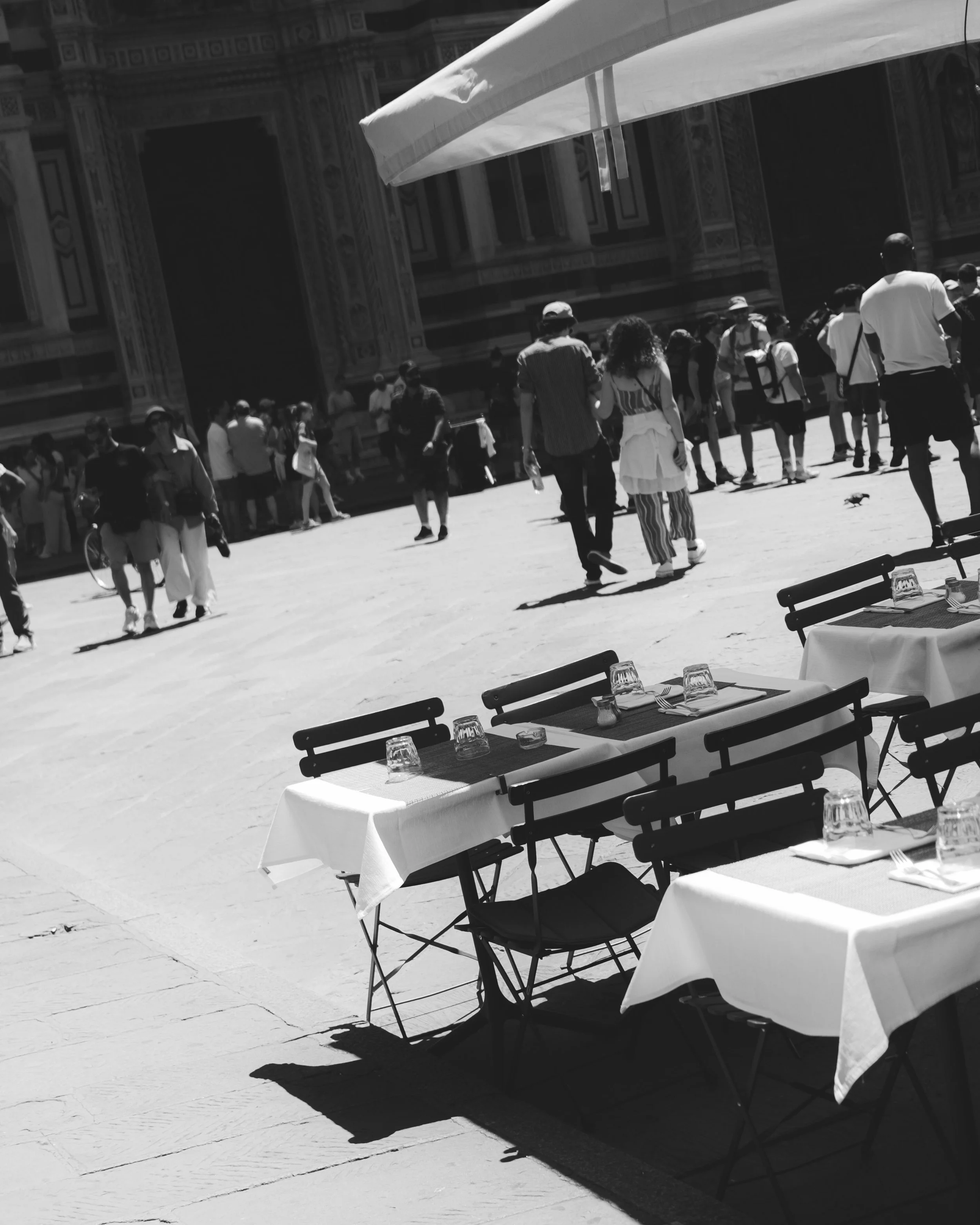 Outdoor terrace with tables and chairs, set with tablecloths, glasses, and utensils, overlooking a busy plaza with pedestrians and historic architecture. Lionel Chu - Hotel & Travel Photographer