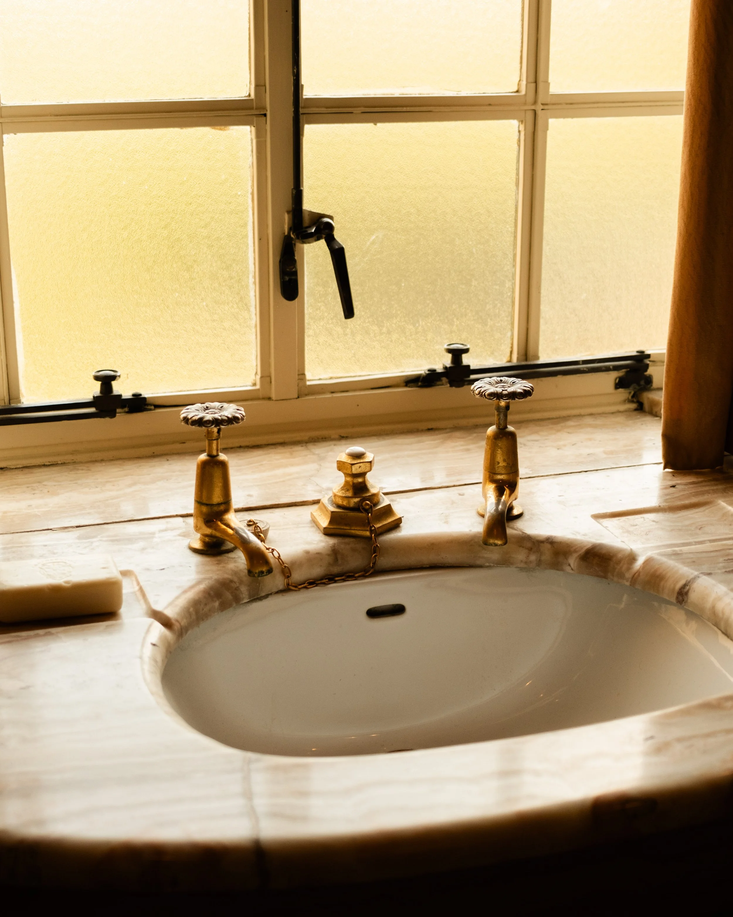 A bathroom sink with vintage brass fixtures, a marble countertop, and a window behind it. The window has closed frosted glass panes and a black handle, with a brown curtain partially visible on the right. Lionel Chu - Hotel & Travel Photographer