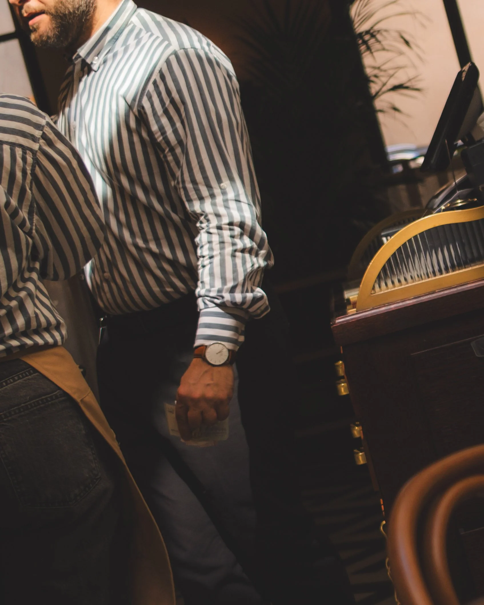 Close-up of two men wearing striped shirts, one with a wristwatch, standing in a dimly lit restaurant or cafe near a piano with a gold trim.