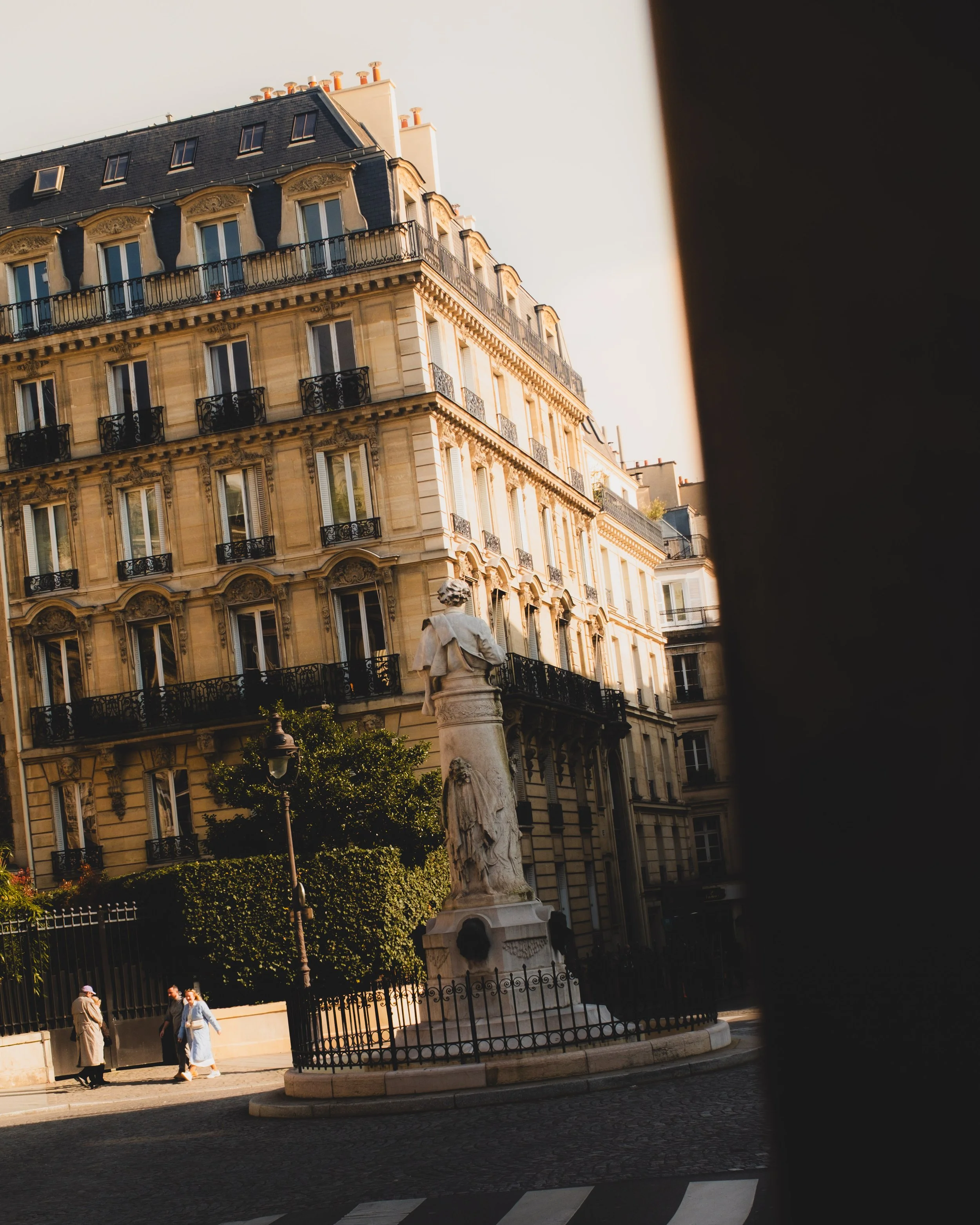 Street view of an elegant Parisian building with ornate balconies and decorative features, featuring a statue on a pedestal surrounded by an iron fence, with pedestrians and a cobblestone street in the foreground. Lionel Chu - Hotel & Travel Photogra