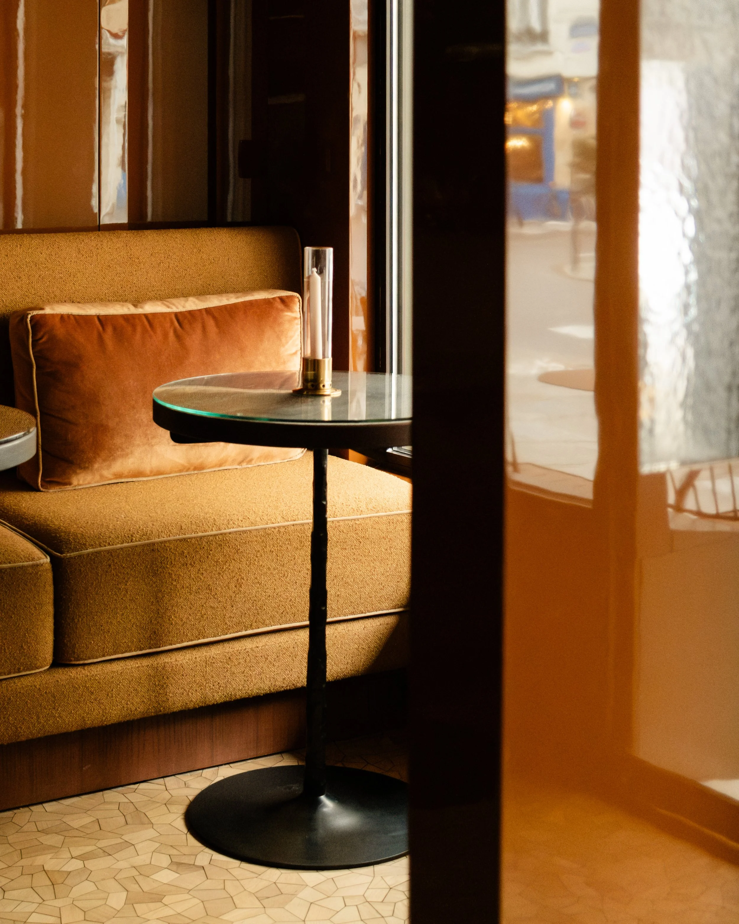 A cozy seating area inside a restaurant or cafe featuring a mustard-colored sofa with a matching pillow, a round glass-top table with a cylindrical candle holder, and a window allowing natural light to illuminate the scene. Lionel Chu