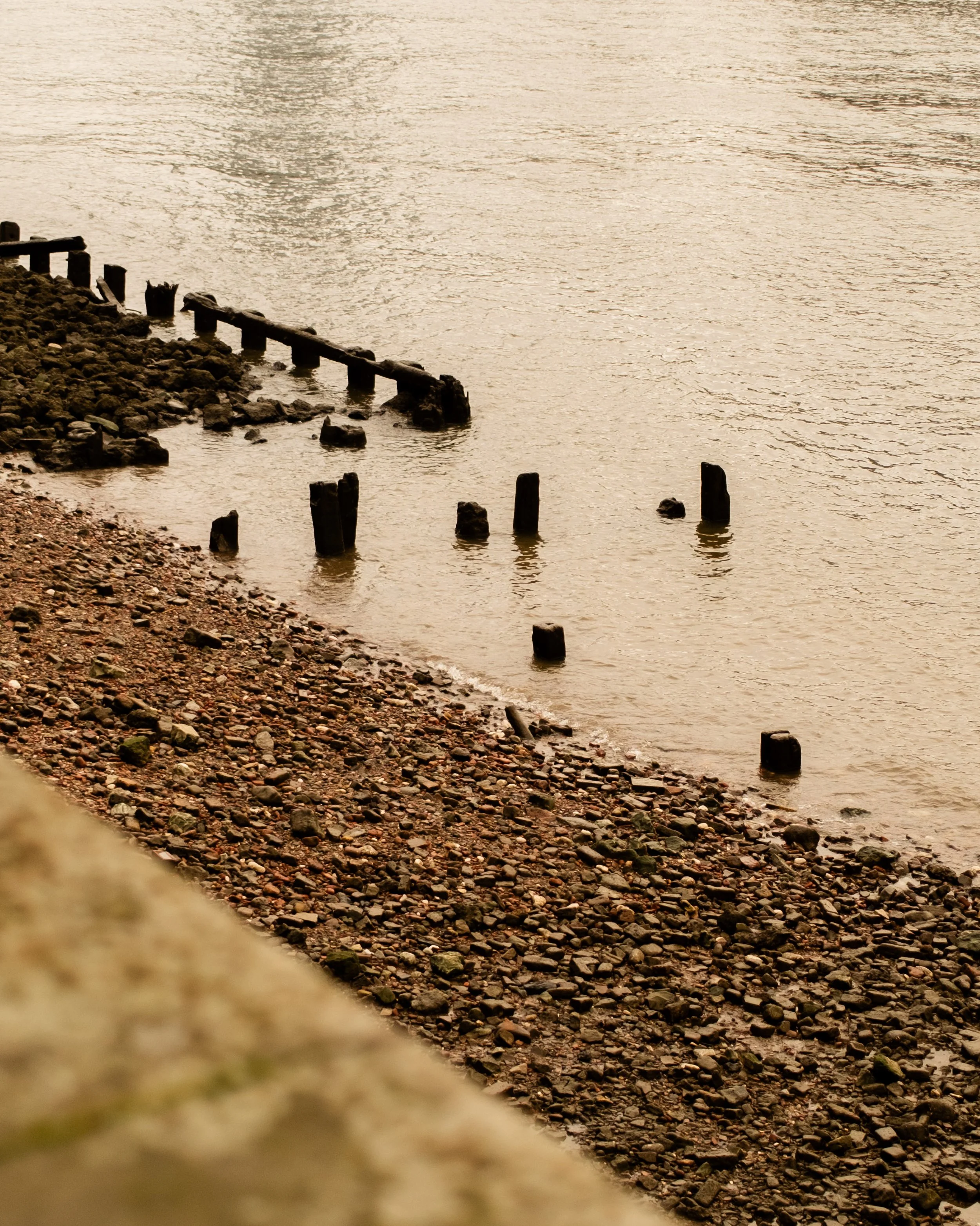 A rocky shoreline with weathered wooden posts extending into a body of water, possibly a lake or ocean, during a calm, overcast day. Lionel Chu - Hotel & Travel Photographer