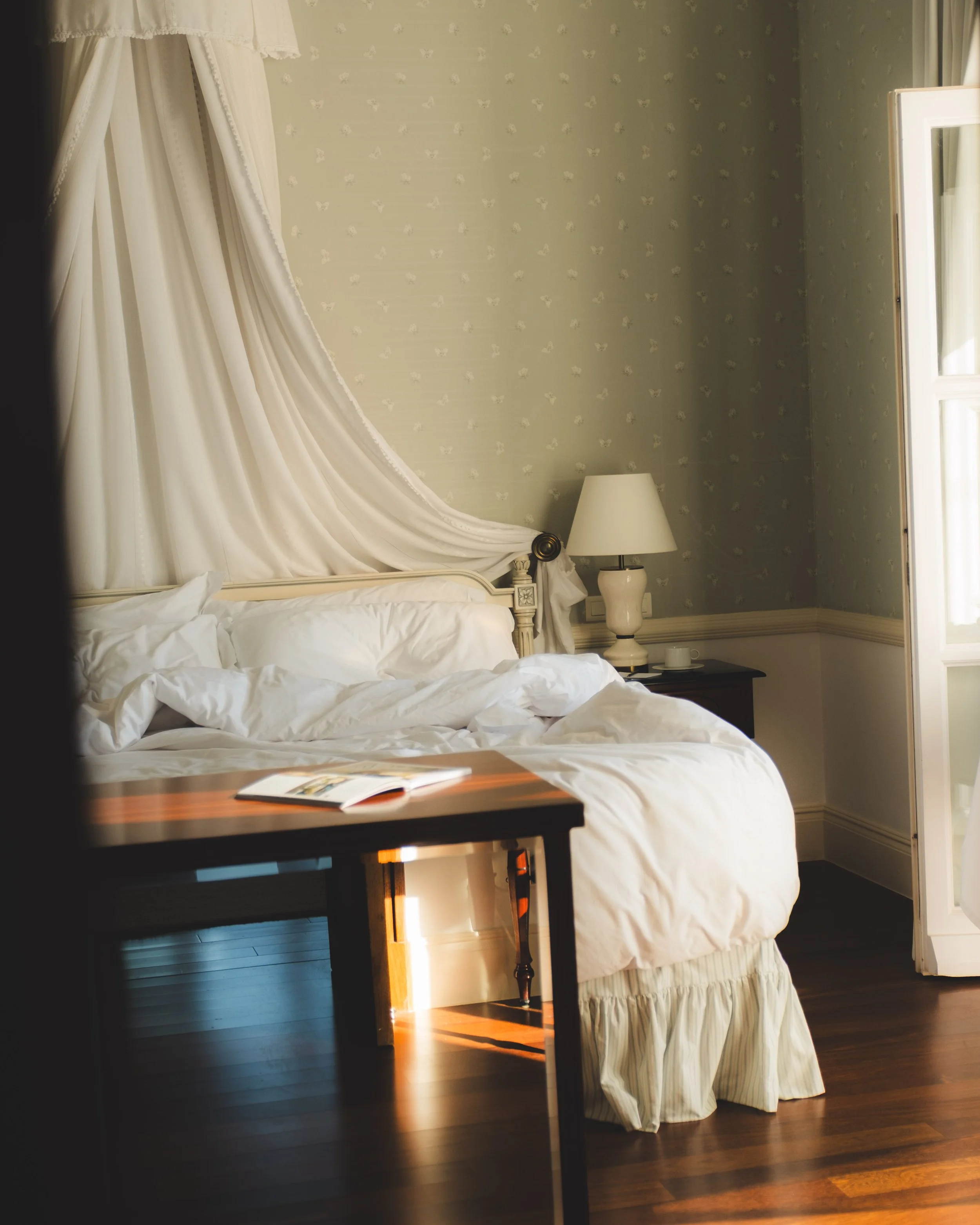 A bedroom with a bed having white linens, decorative pillows, and a canopy with cream-colored fabric. There is a nightstand with a white lamp, and a window with cream curtains. The room has vintage-style wallpaper with small white floral patterns on 