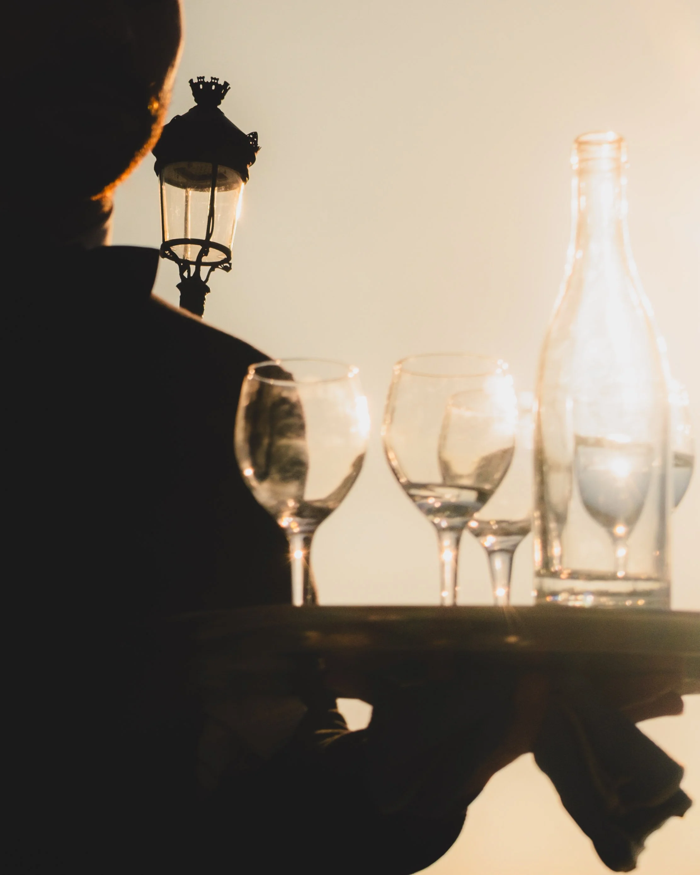 Silhouette of a waiter holding a tray with empty wine glasses and a bottle, with a lamp post in the background at sunset. Lionel Chu - Hotel & Travel Photographer.