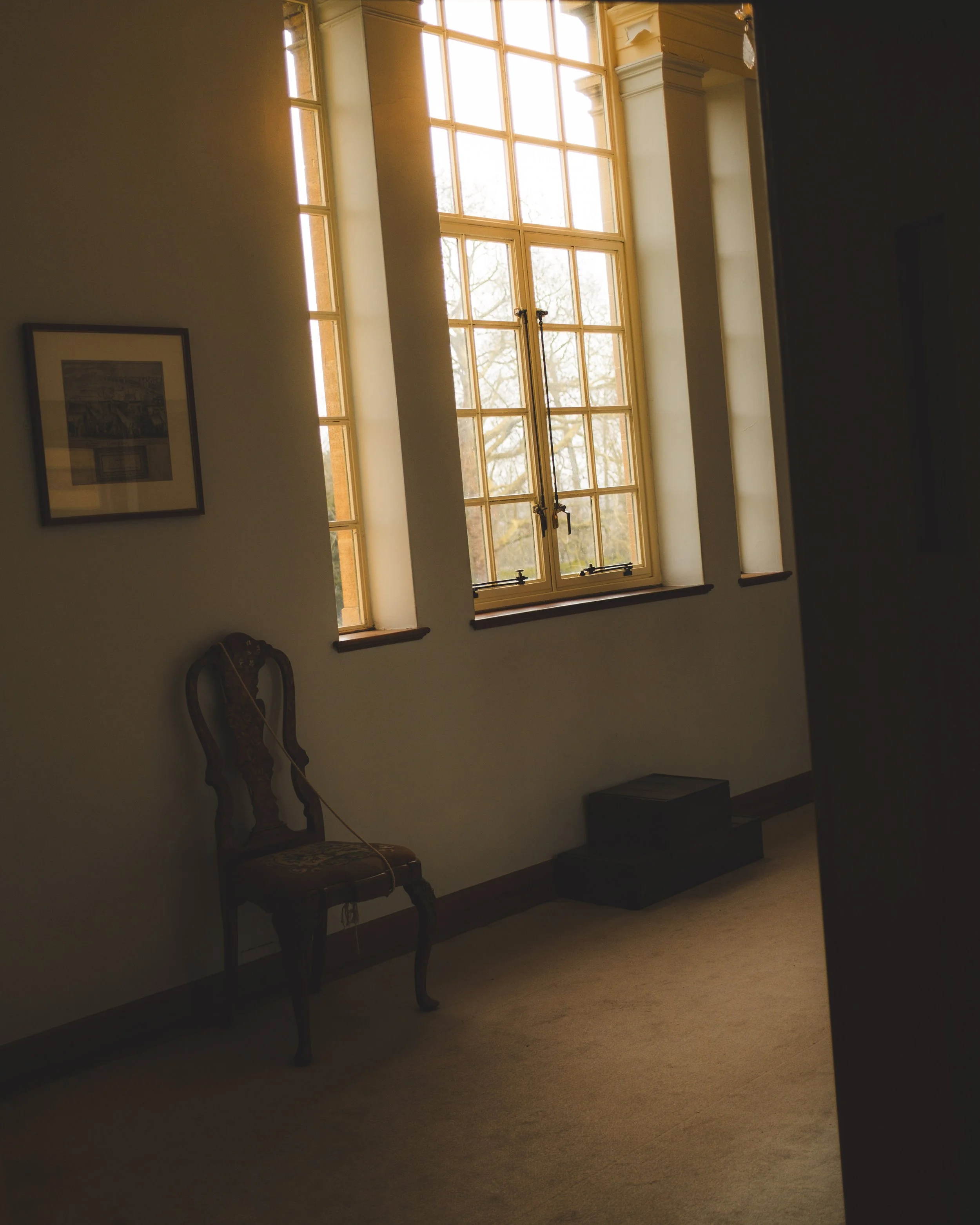 A large window with sunlight streaming through, framing a view of bare tree branches outside. The interior includes a wall with a framed picture, an elegant chair, and a black bench beside a beige carpet.