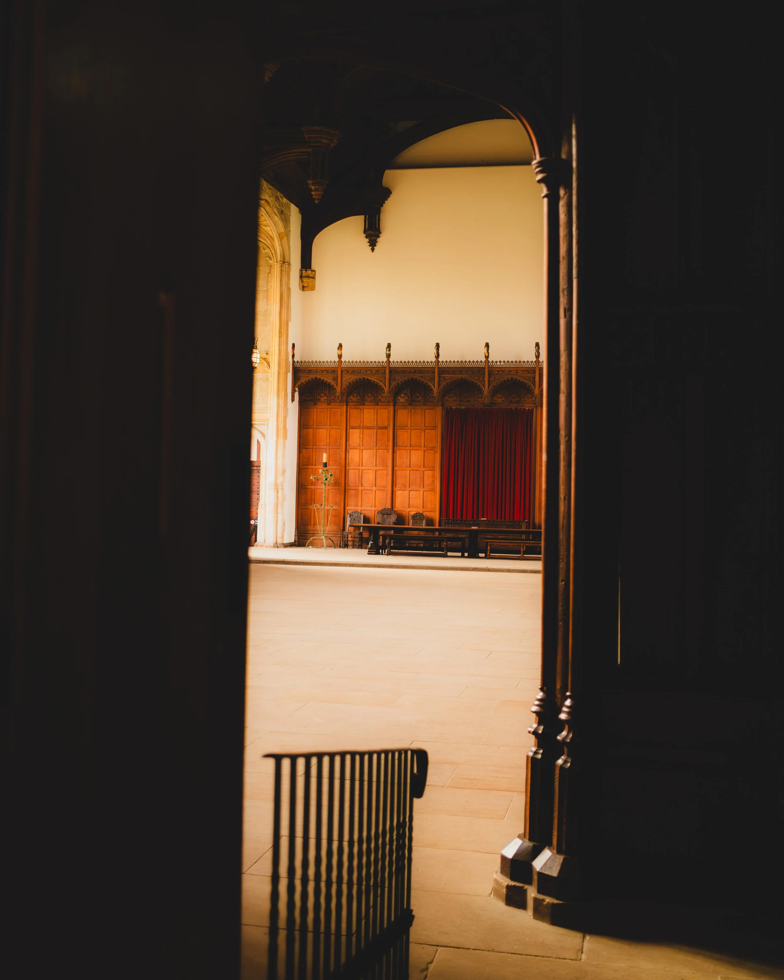 An interior view of a large room, possibly a hall or auditorium, as seen through a partially opened dark wooden door. The room features wooden paneling on the walls, a red curtain, some benches, and a decorative candelabrum, with a high ceiling suppo