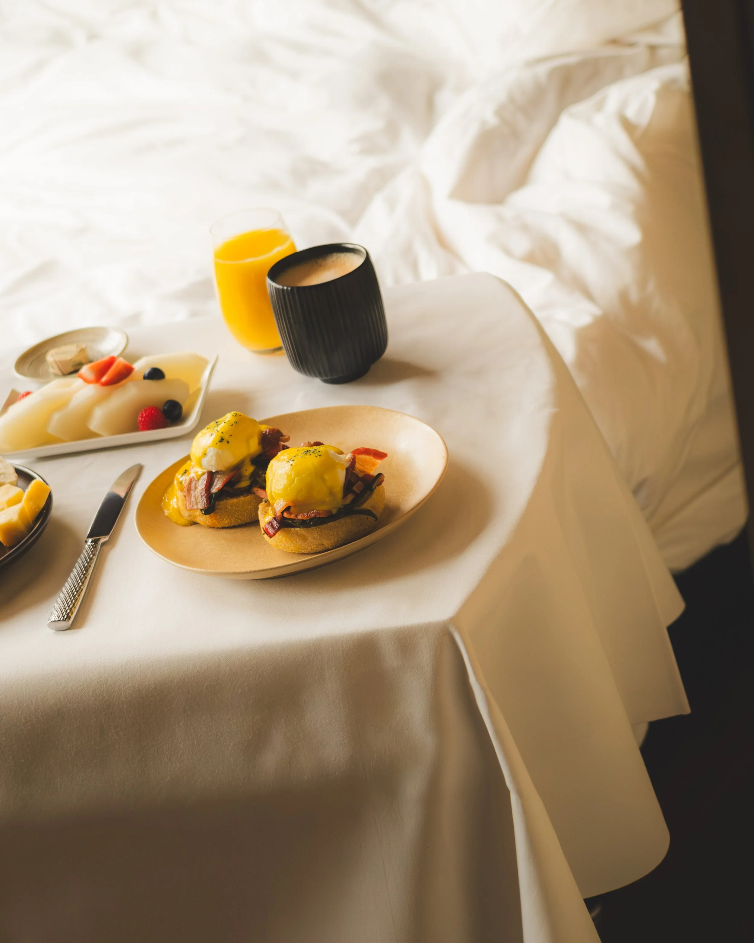 A table set with plates of eggs Benedict, a glass of orange juice, and a cup of coffee, with a bed in the background.