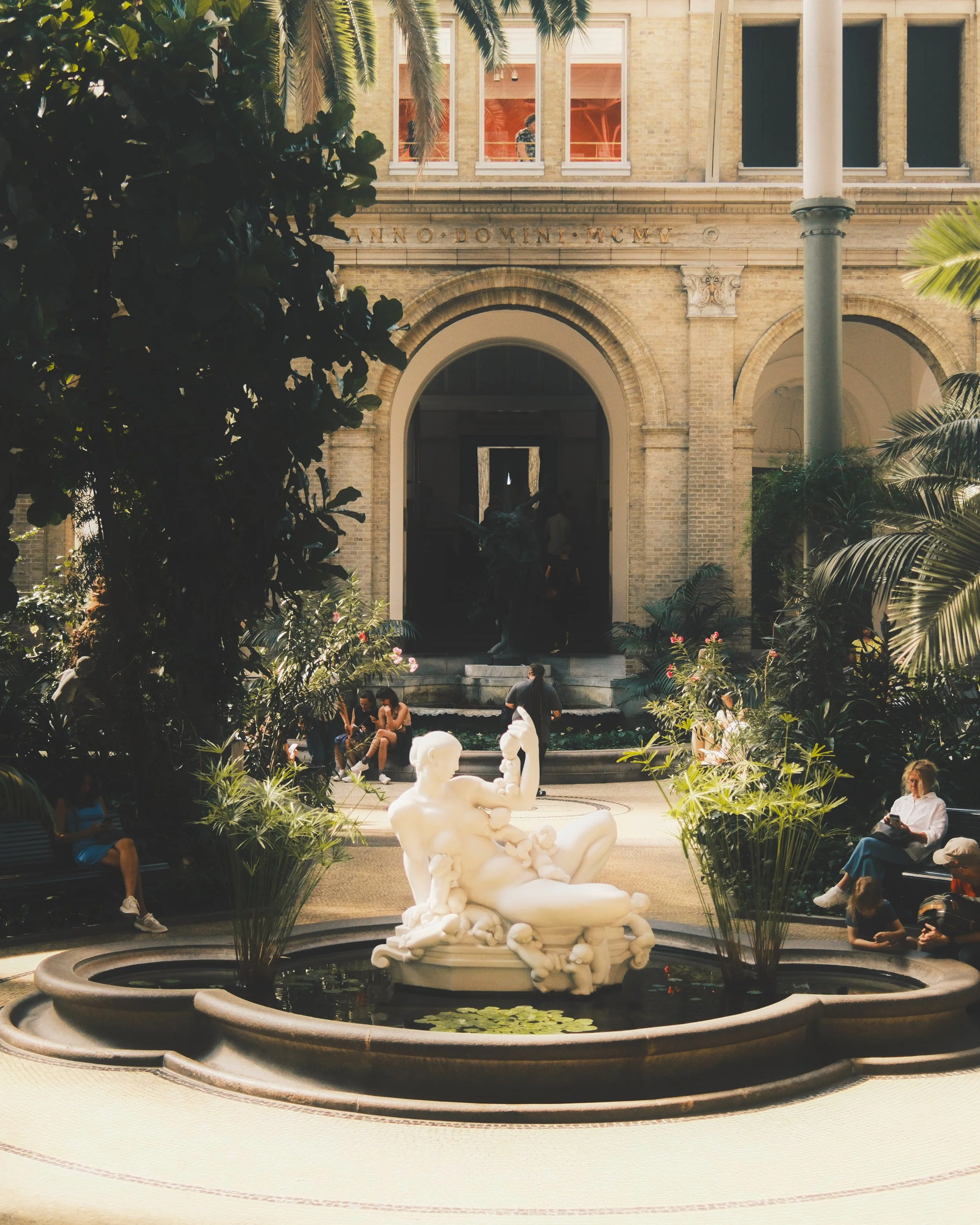 Indoor courtyard with a white sculpture at the center of a fountain, surrounded by greenery and people sitting on benches. The background shows an arched entrance to a building with visible architectural details. Ny Carlsberg Glyptotek, Copenhagen.