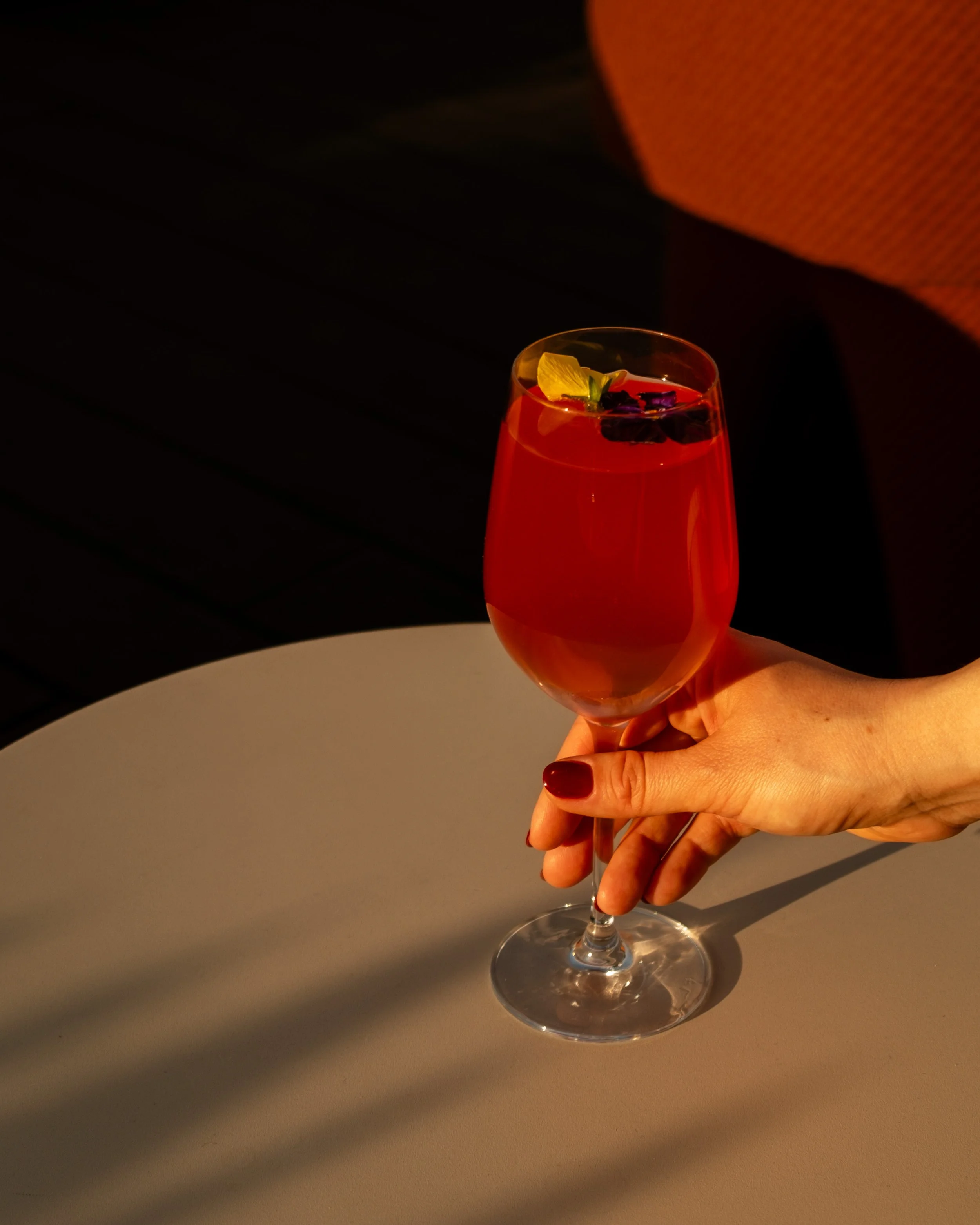 Hand holding a glass of red cocktail with fruit garnish, on a round white table with dark background. Lionel Chu - Hotel & Travel Photographer
