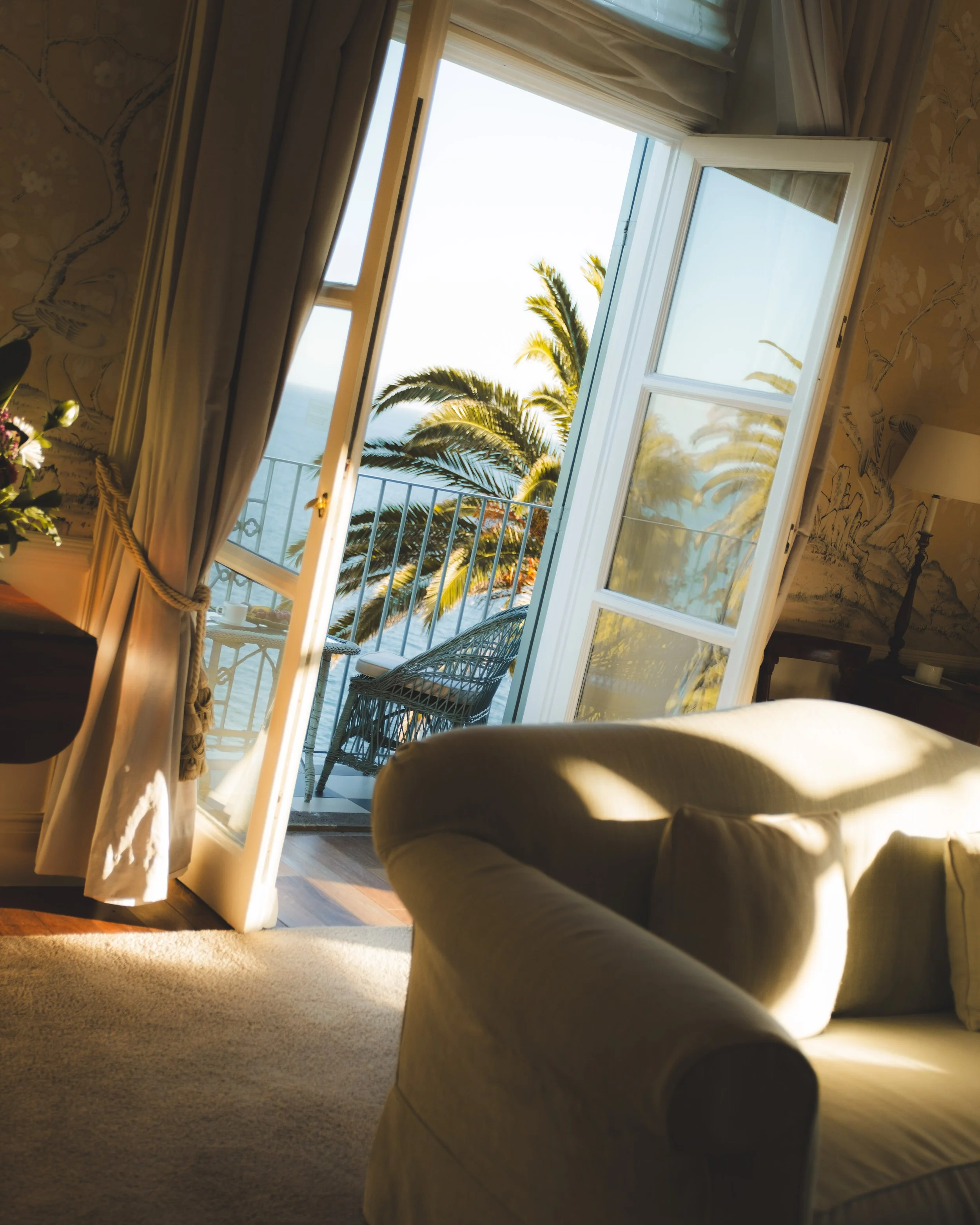 A room with an open glass door leading to a balcony, revealing palm trees and a view of the ocean. Inside the room, there is beige carpet, a beige armchair, and floral wallpaper.