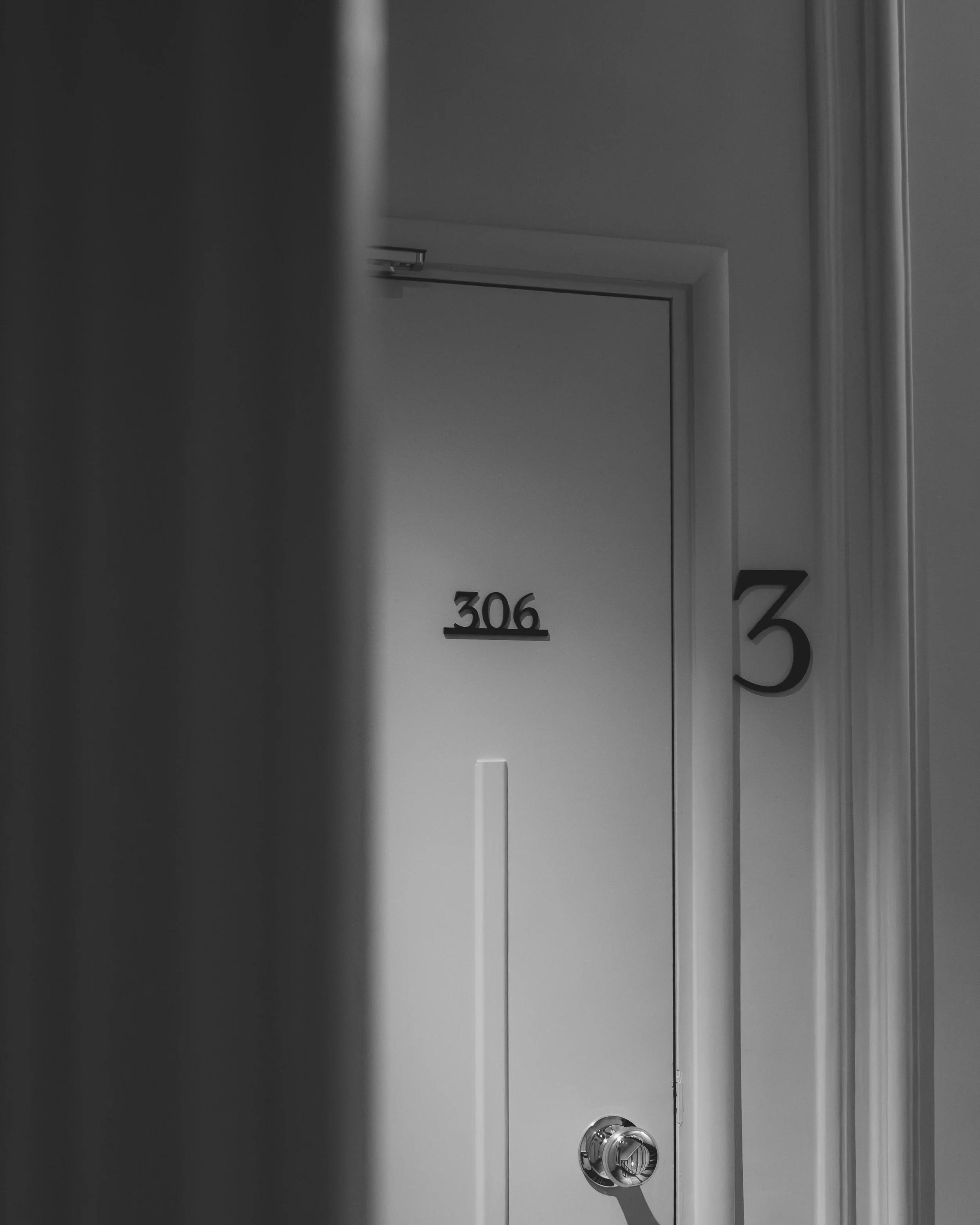 A white door with the room number 306 on it, a door handle, and a lock. The door frame and wall are white, with some shadows visible.