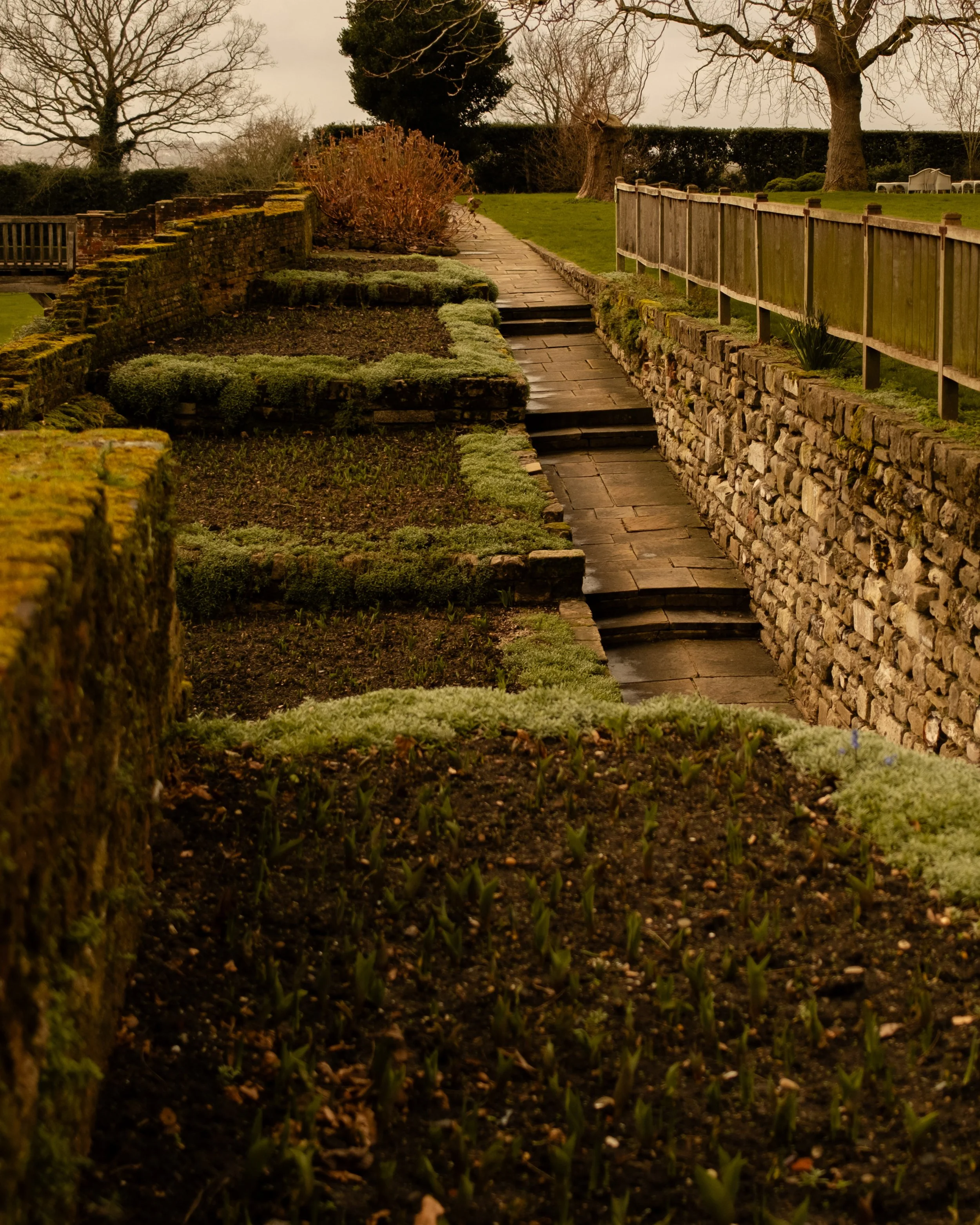 A stepped stone pathway runs through a landscaped garden with trees, bushes, and a moss-covered stone wall on the right side. The sky is overcast. Lionel Chu - Hotel & Travel Photographer