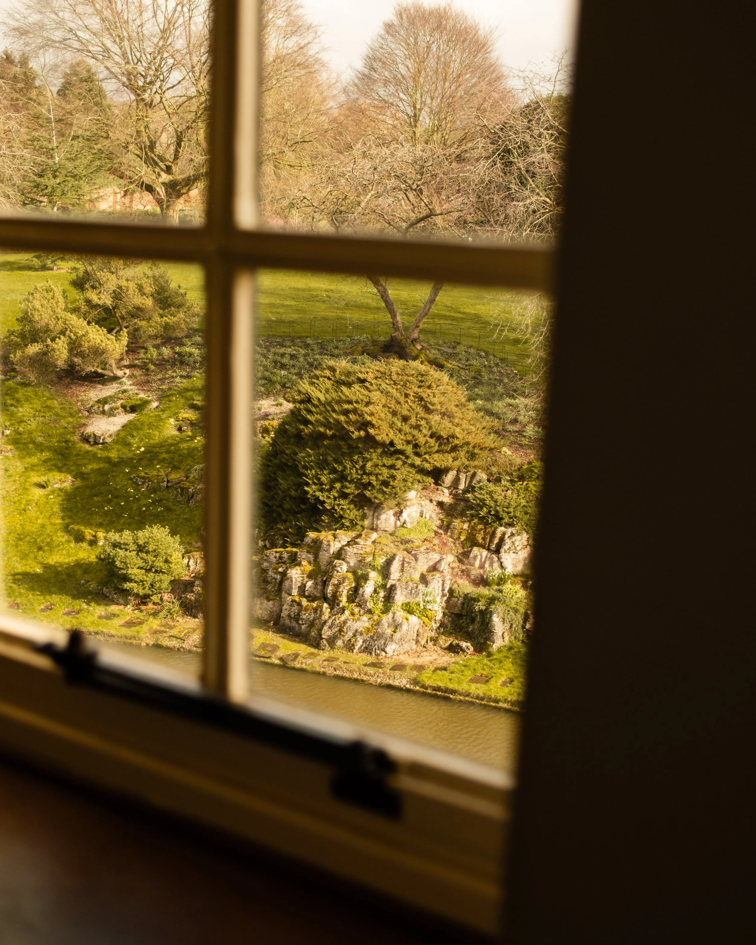 View of a garden with trees, shrubs, rocks, and a body of water seen through a window with divided panes. Lionel Chu - Hotel & Travel Photographer