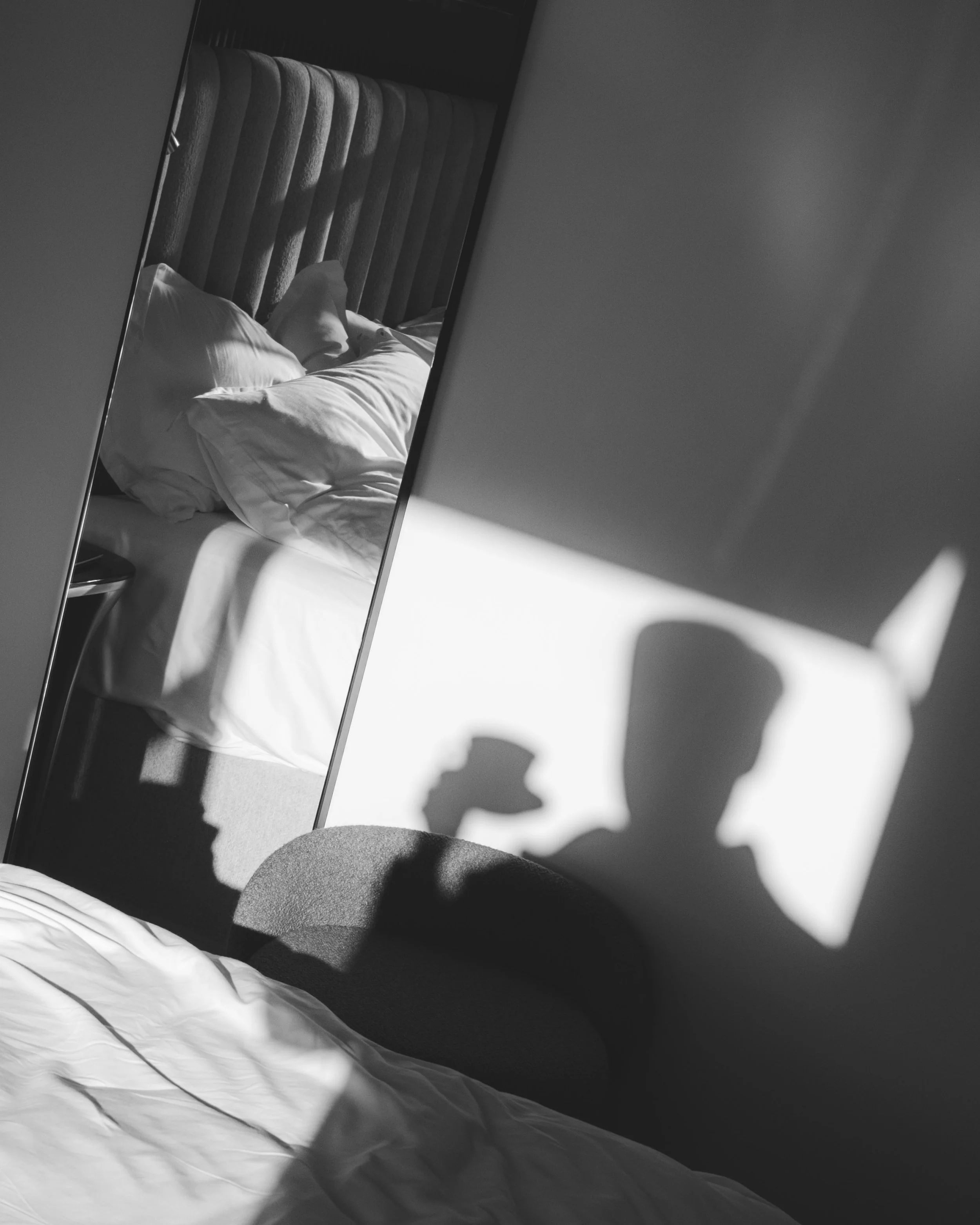 A black and white photo of a bed with white pillows and bedsheets. A headboard with vertical stripes is visible at the head of the bed. A mirror on the wall reflects the bed and part of the headboard. Shadows are cast on the bedsheets and wall.