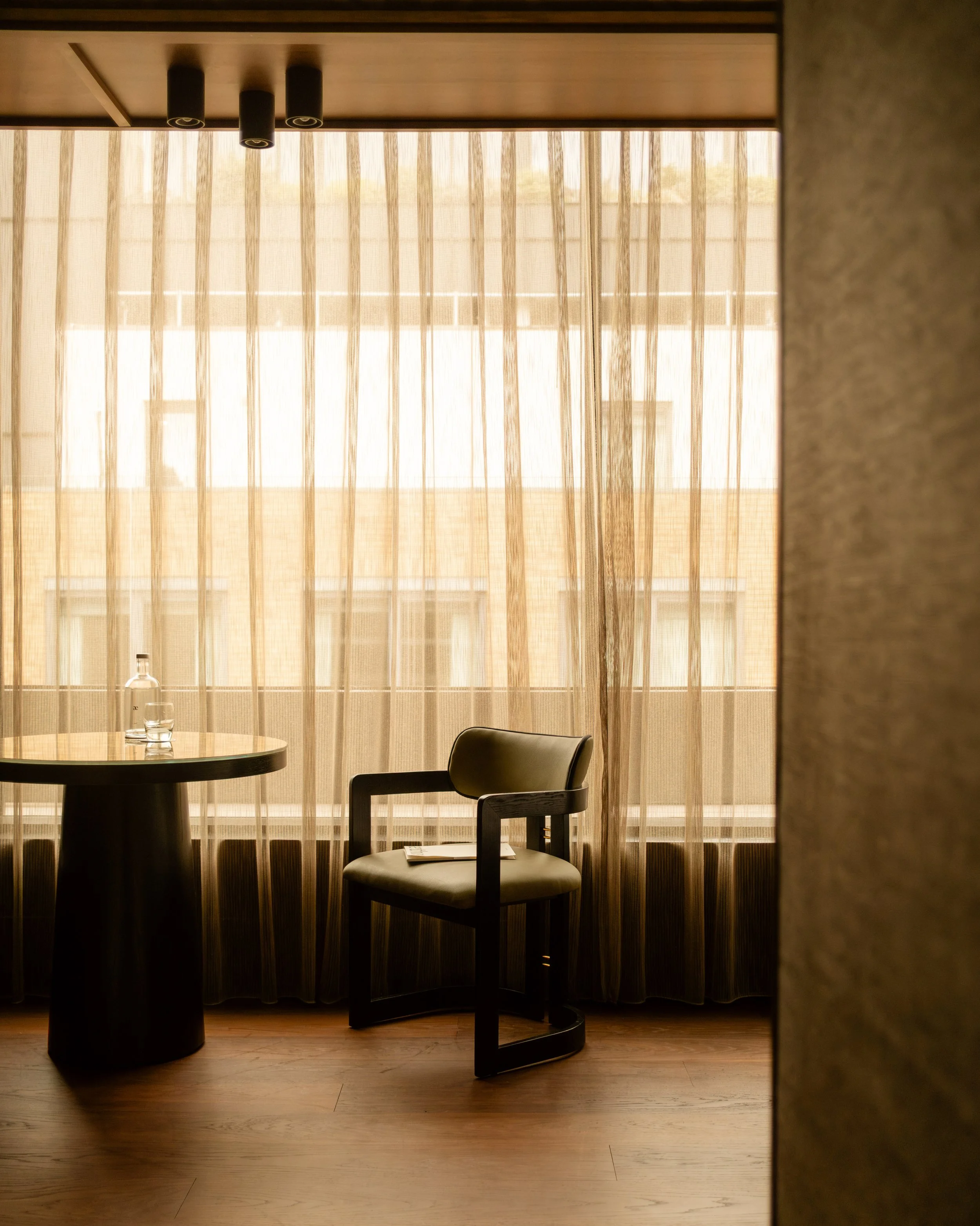Interior view of a hotel room with a window covered by beige curtains. A small table with a bottle of water and a glass, and a black armchair with a beige cushion, are near the window. Lionel Chu - Hotel & Travel Photographer