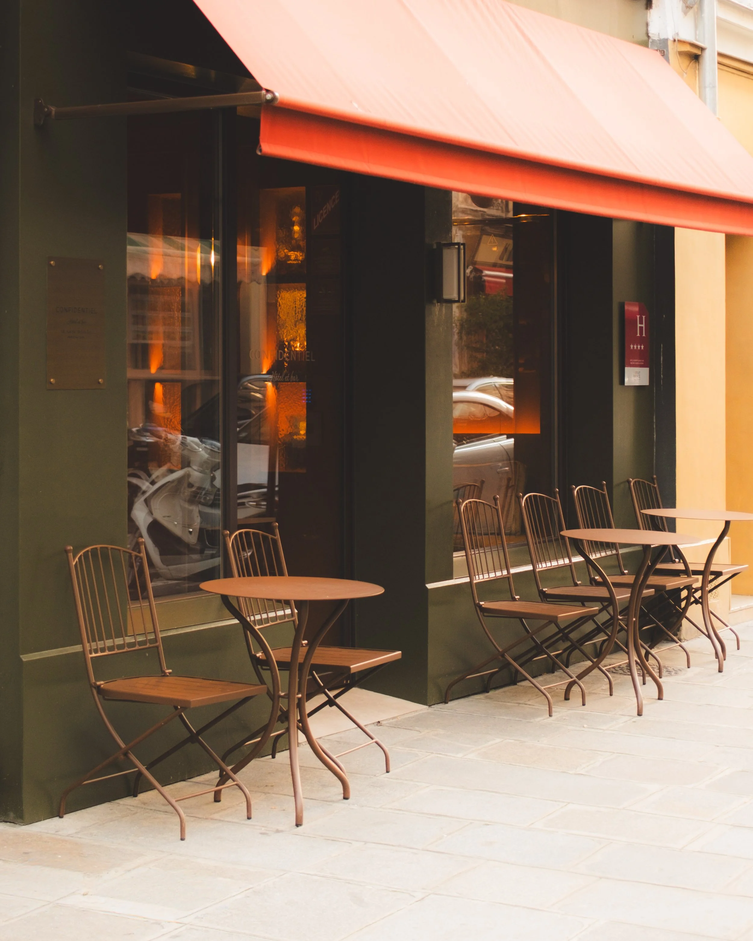 Outside cafe seating with three wicker chairs and round table on a stone patio, with a green wall and large glass windows.