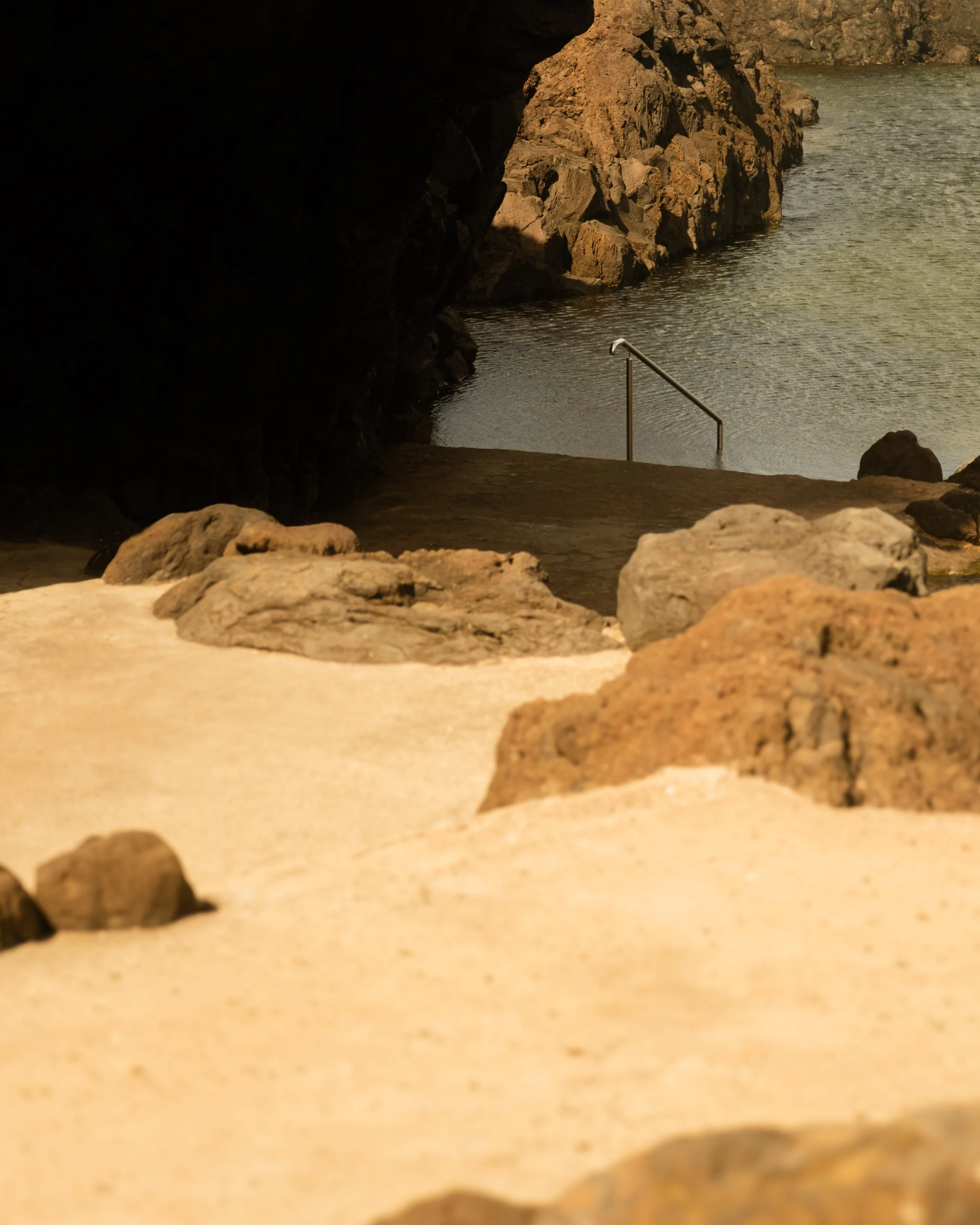 View from a beach looking out towards a rocky shoreline with water, a metal railing extends into the water, and rocks are scattered in the foreground.