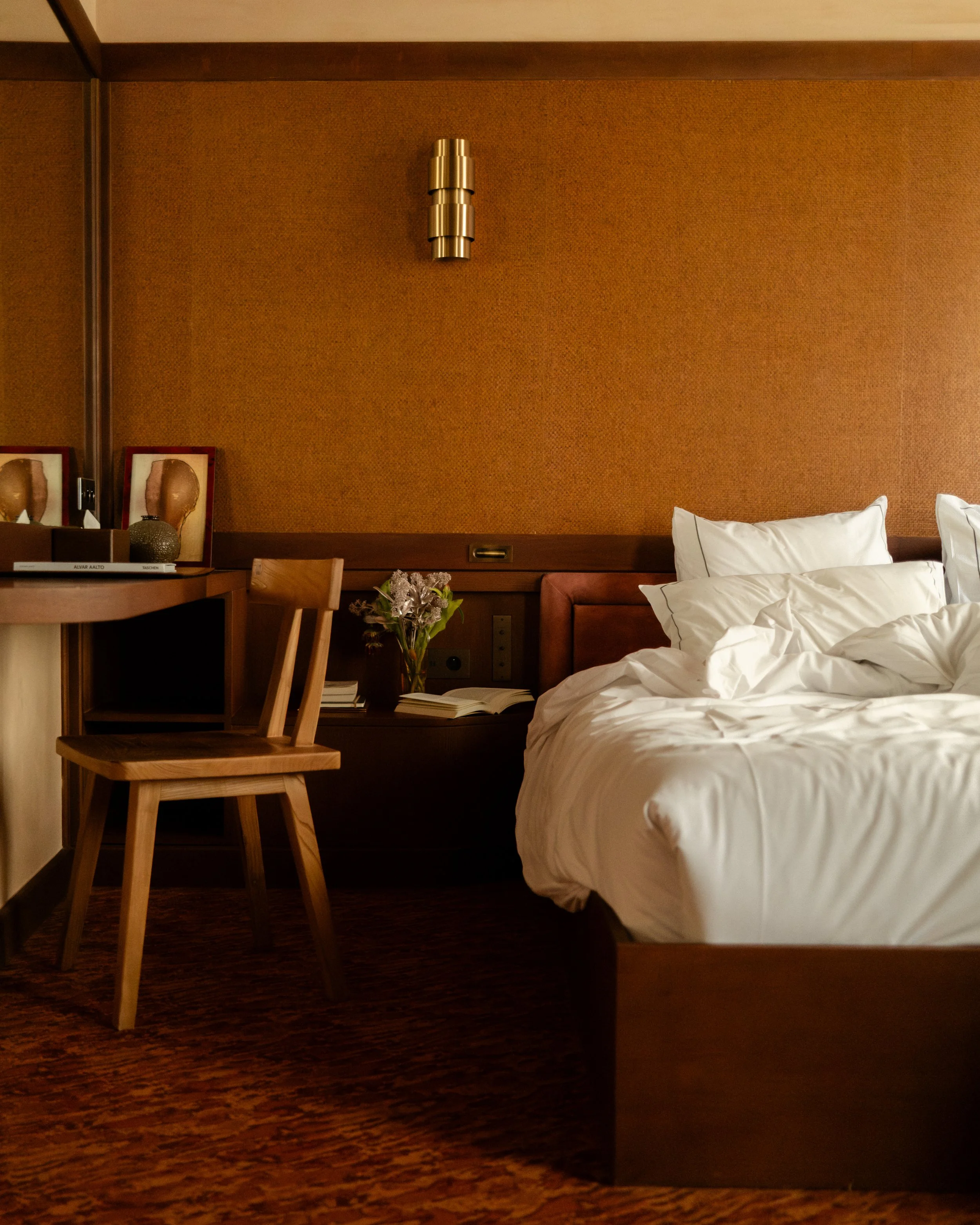 Hotel room with a made bed, white pillows, a wooden chair, a small table with a vase of flowers, a book, and framed artwork on the wall. Lionel Chu - Hotel & Travel Photography