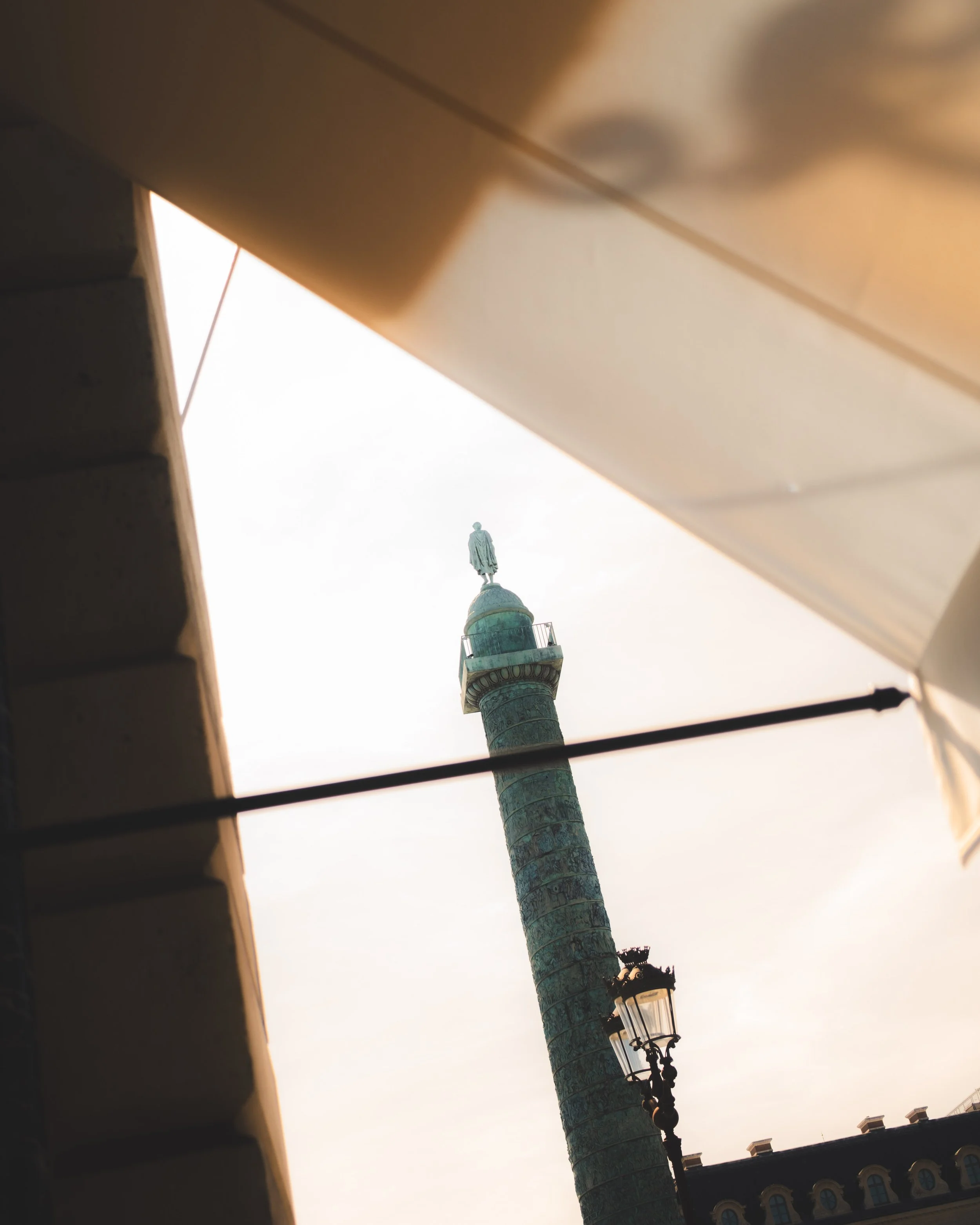 View of a tall monument with a statue at the top, seen from below, framed by two buildings with awnings and a street lamp.