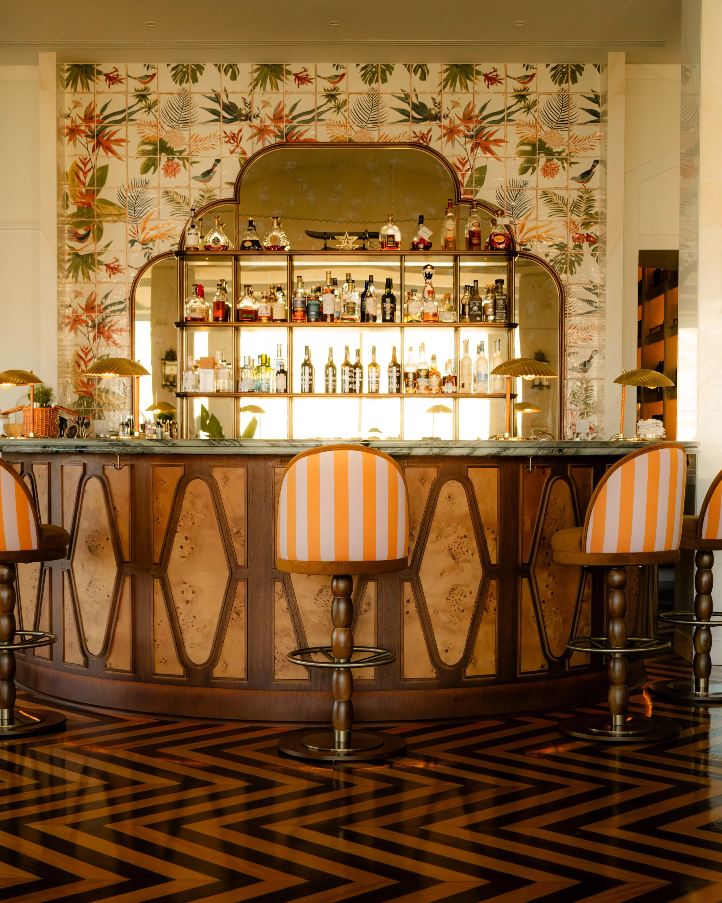 Bar with a variety of liquor bottles on shelves, decorative wallpaper with bird and leaf patterns, wooden bar counter, striped chairs, and a checkered wooden floor.