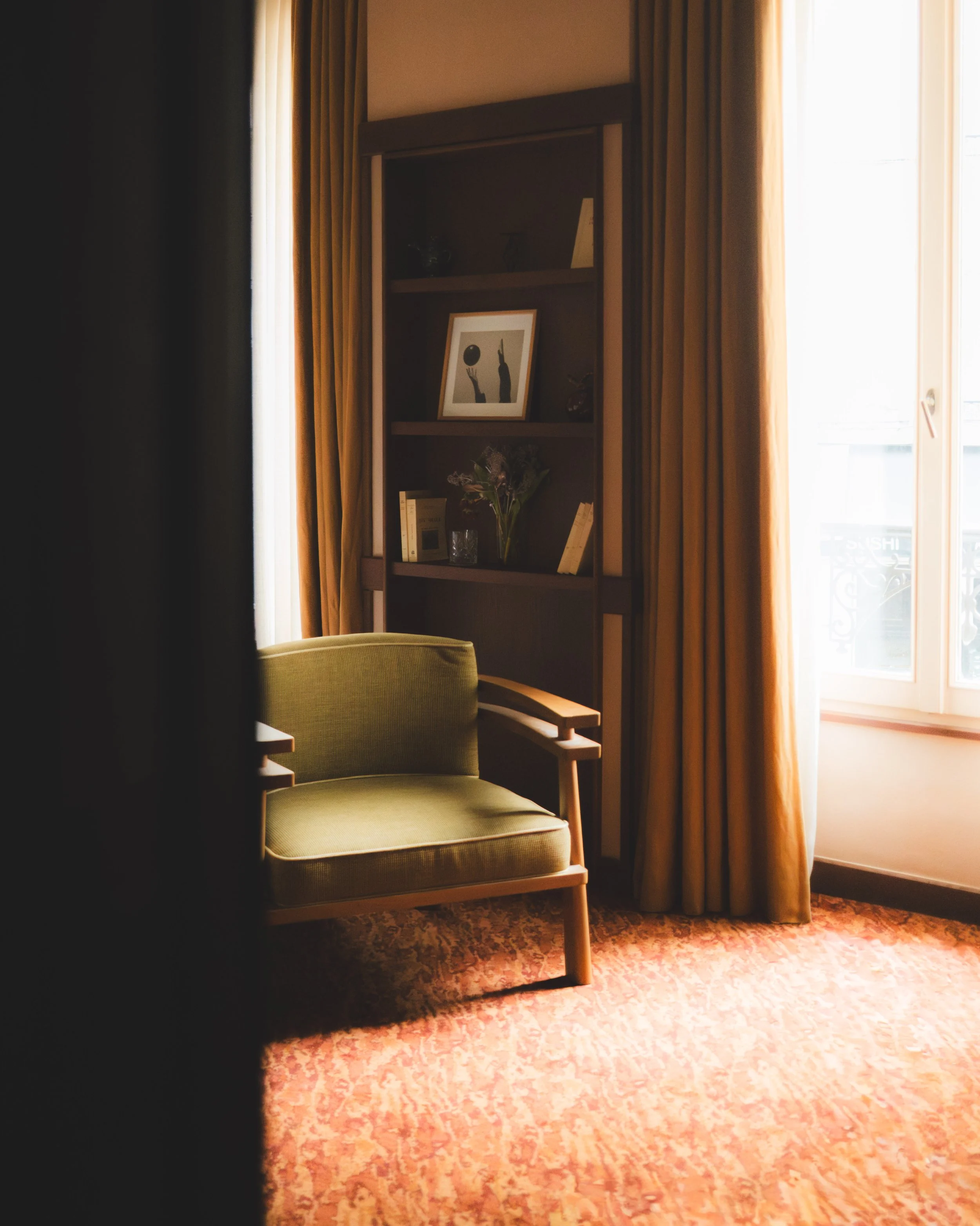 A corner of a room with a green upholstered armchair, a tall shelf with decorative items, a framed black-and-white artwork of two hands reaching for a ball, and an open door with a window letting in natural light. The room has beige walls, gold curtains, and a patterned carpet.
