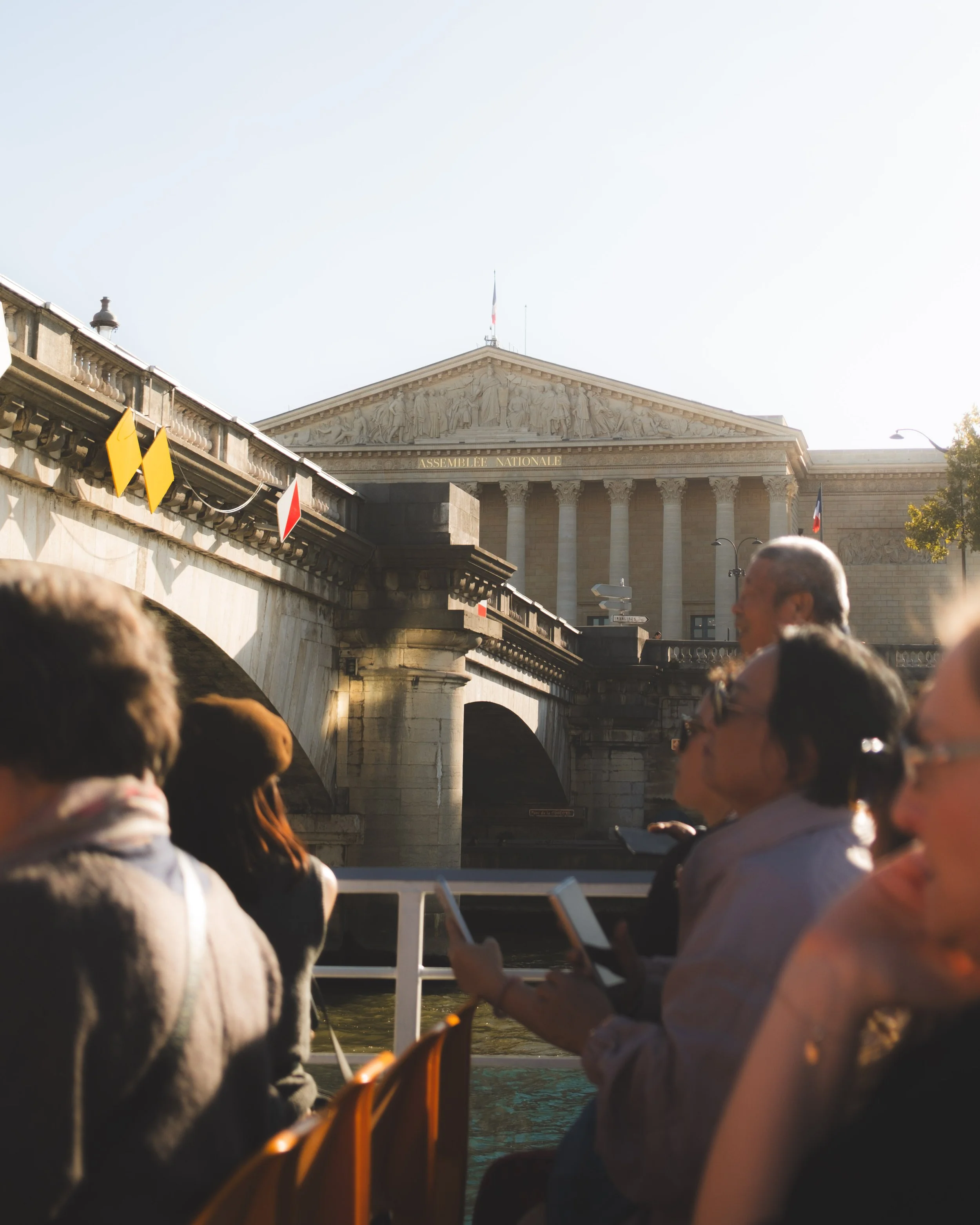 A crowd of people sitting by a river, with a bridge featuring multiple arches and classical architecture in the background, including a building with columns and a pediment with statues and the words 'Assemblée Nationale'. Some people are using elect