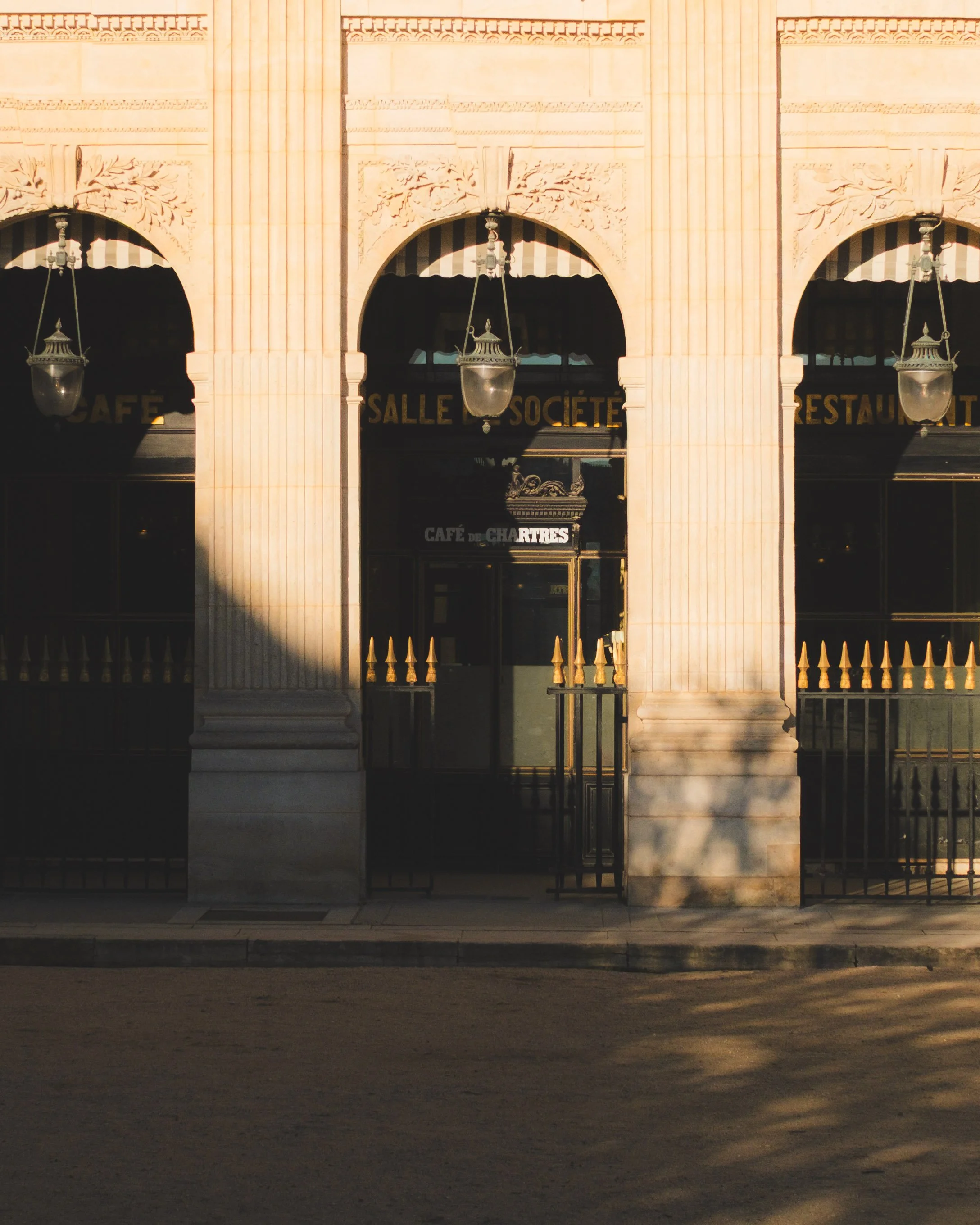 Exterior of a building with three arched entrances, ornate carvings, and hanging lanterns, with signs indicating a café and restaurant in French, illuminated by warm light at dusk.