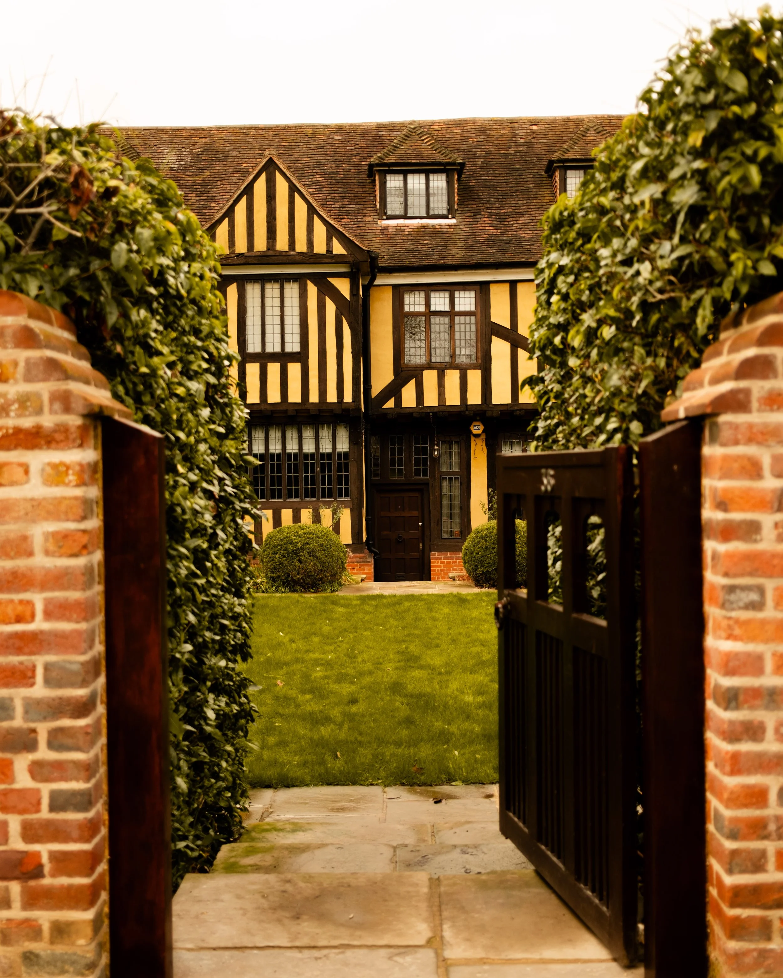 A brick gate opens to a grassy lawn leading to a historic, black-and-yellow half-timbered house with multiple windows and shrubs in the front yard. Lionel Chu - Hotel & Travel Photographer