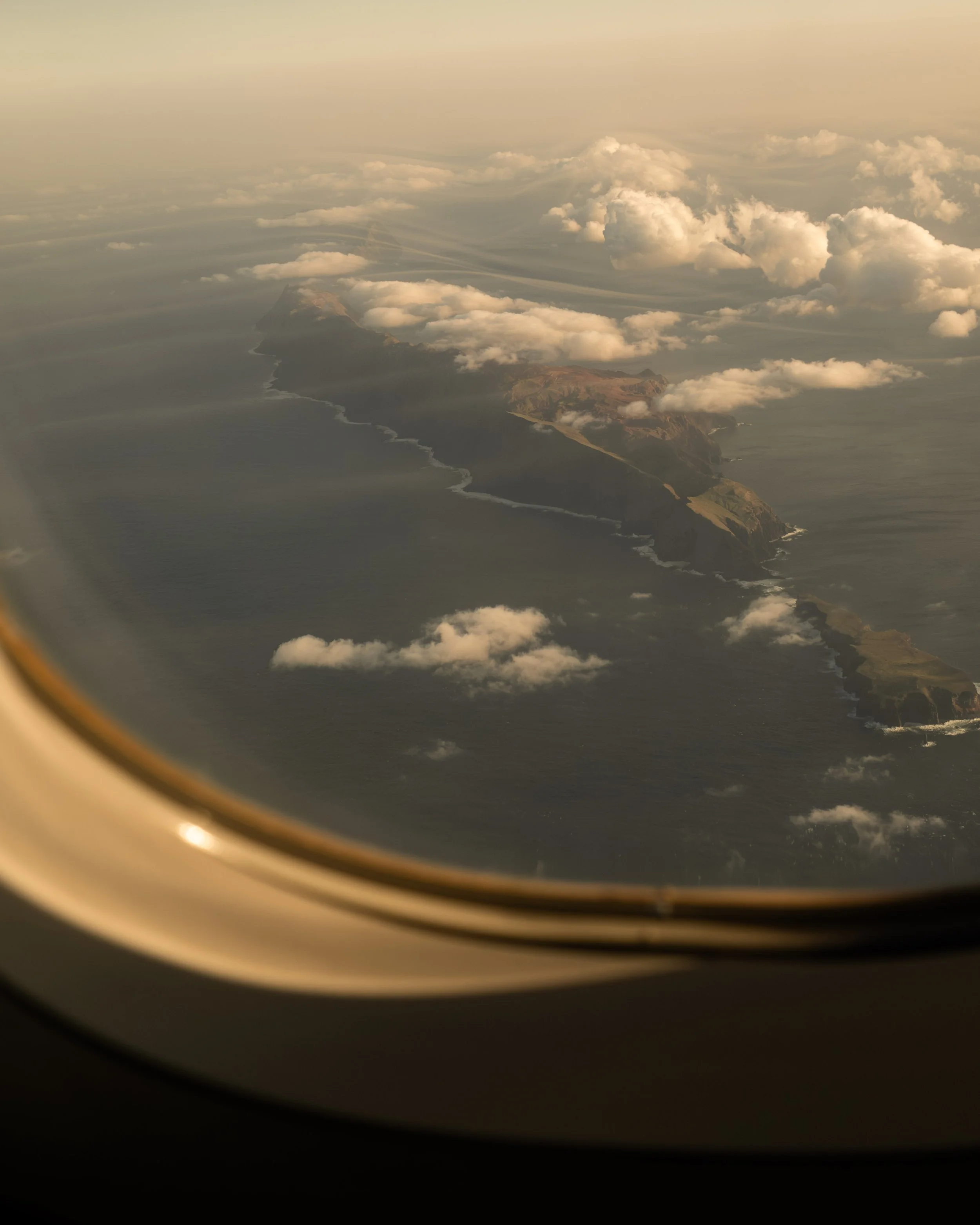 An aerial view of a coastline with rugged cliffs, green patches, and stormy clouds over the sea, seen from an airplane window.