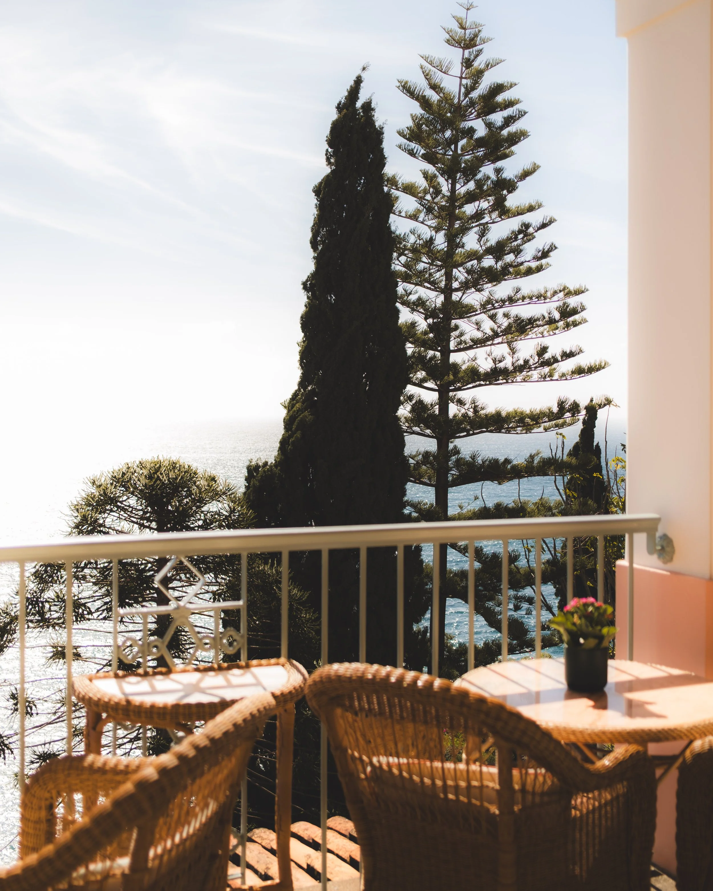 A balcony with a table and wicker chairs overlooking trees and the ocean under a partly cloudy sky.