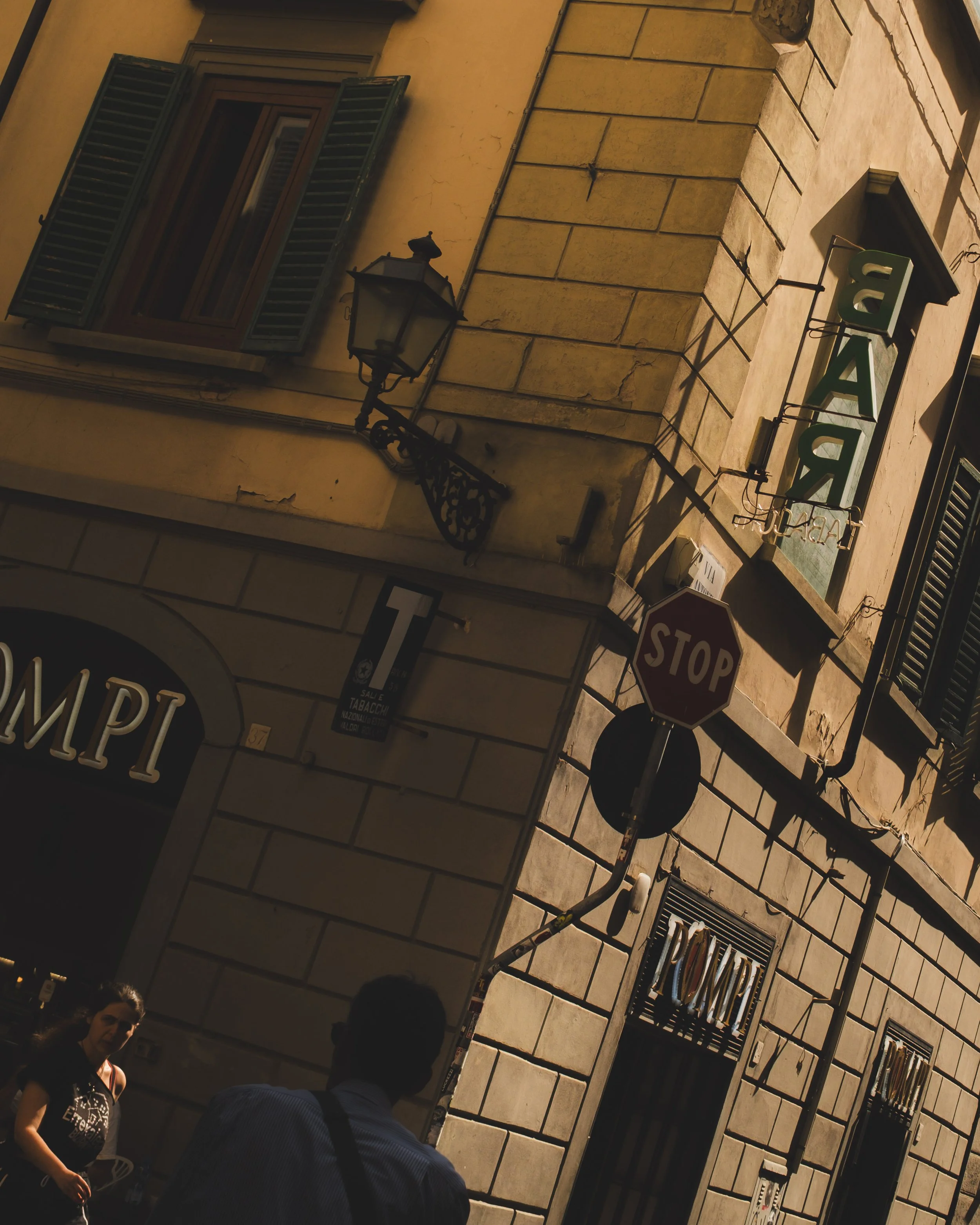 A street corner in Italy with a building featuring green shutters, a street lamp, a stop sign, and a neon sign that says 'BAR'. There are two people in the foreground, one woman and one man, near a shop with a sign written in Italian. Lionel Chu - Ho