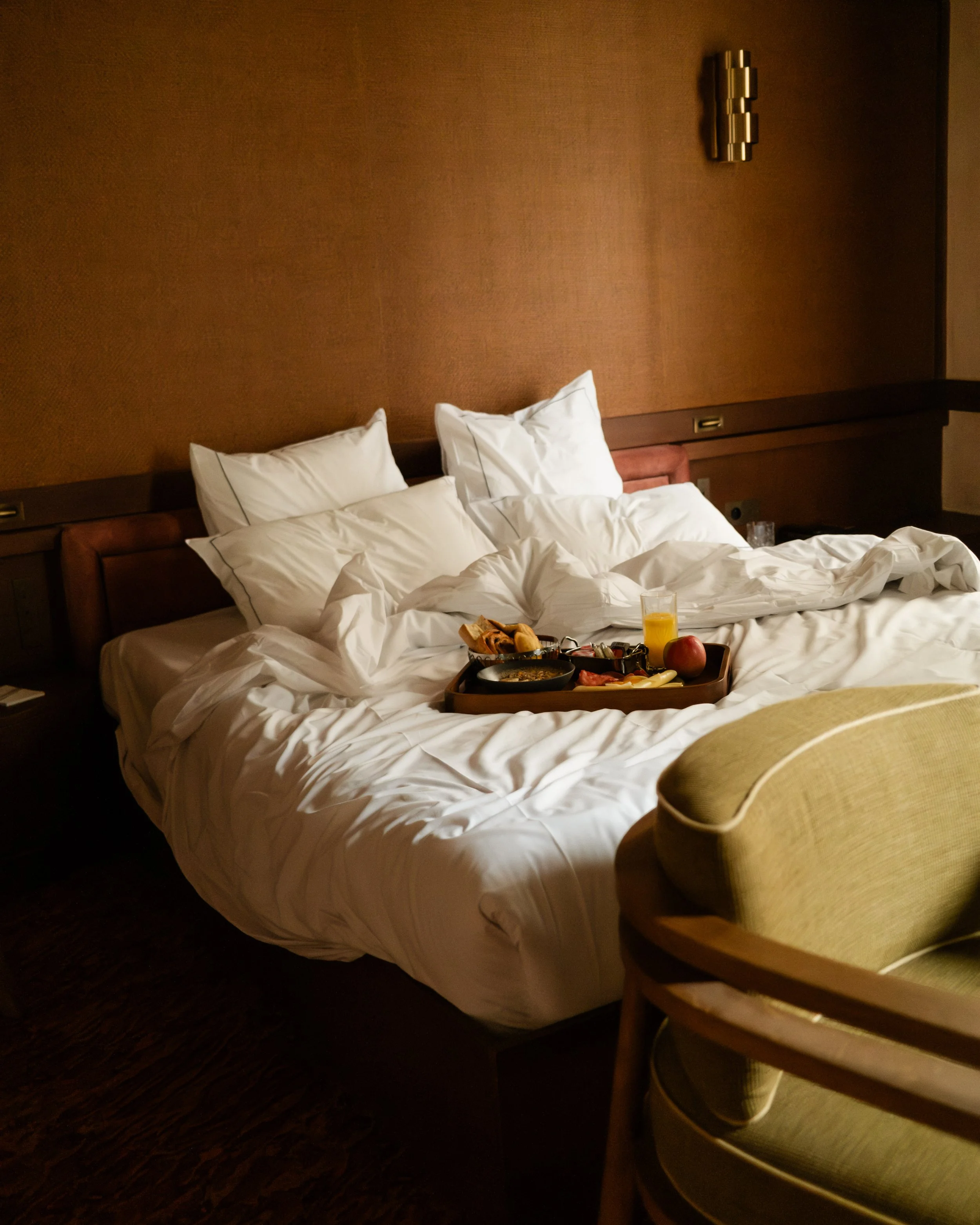 Unmade hotel bed with a breakfast tray, a glass of orange juice, an apple, and breakfast items, in a dimly lit hotel room. Lionel Chu - Hotel & Travel Photographer