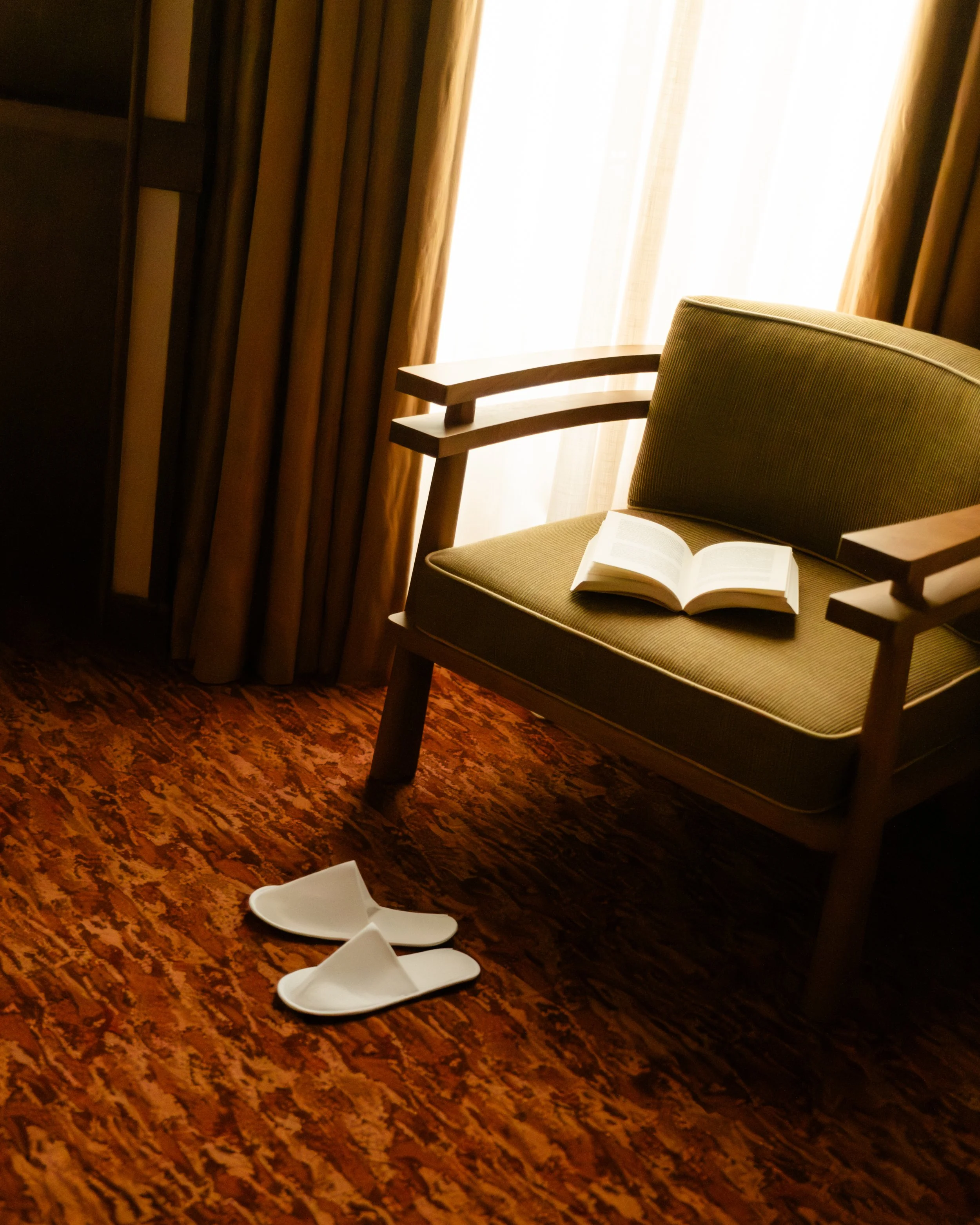 A green armchair with an open book resting on it, positioned near a window with beige curtains, a pair of white slippers on a patterned red-brown carpet. Lionel Chu - Hotel & Travel Photography