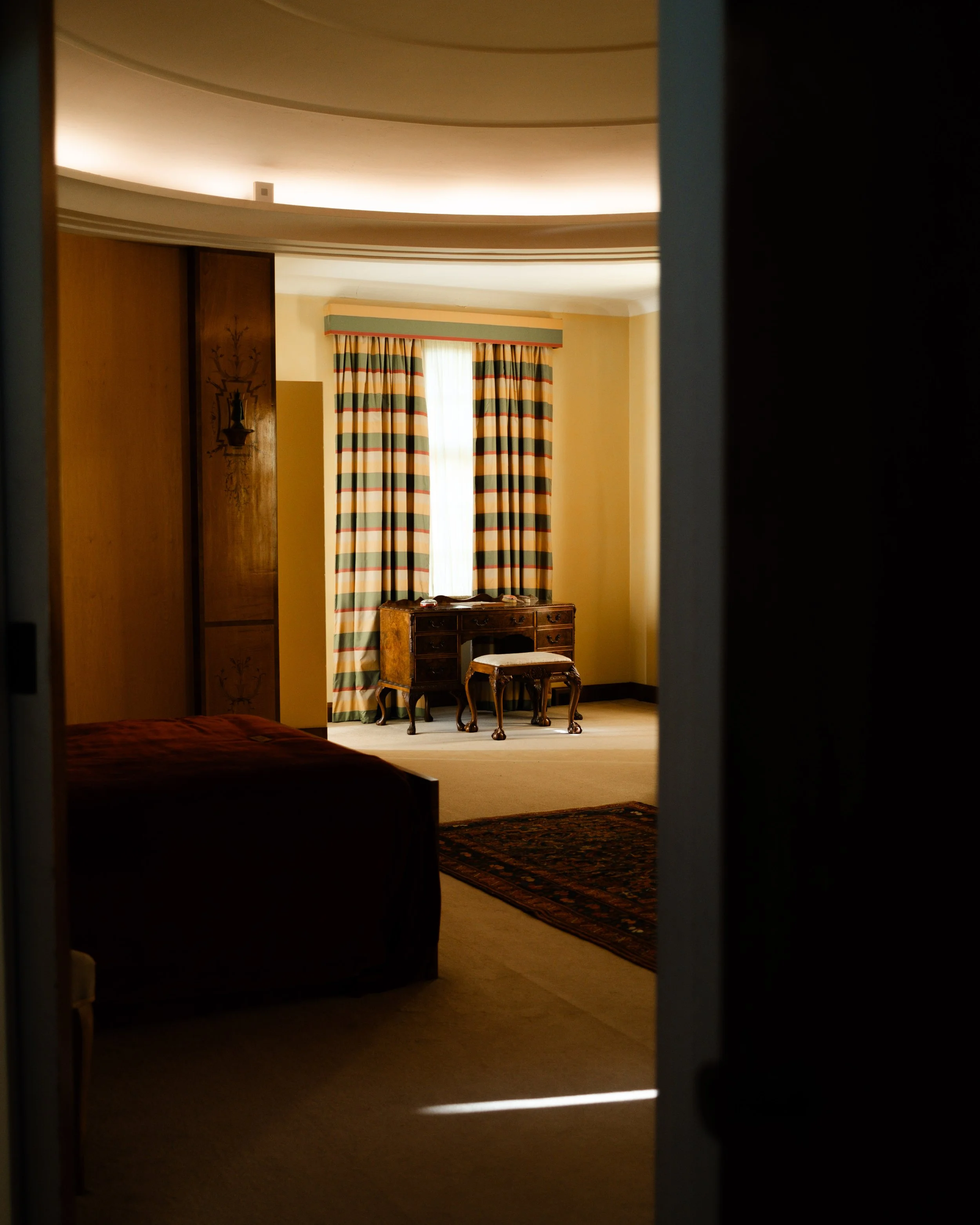 View of a mansion bedroom through a partially open door, showing a bed, a window with striped curtains, a wooden desk with a matching stool, and a decorative rug on the floor. Lionel Chu - Hotel & Travel Photographer