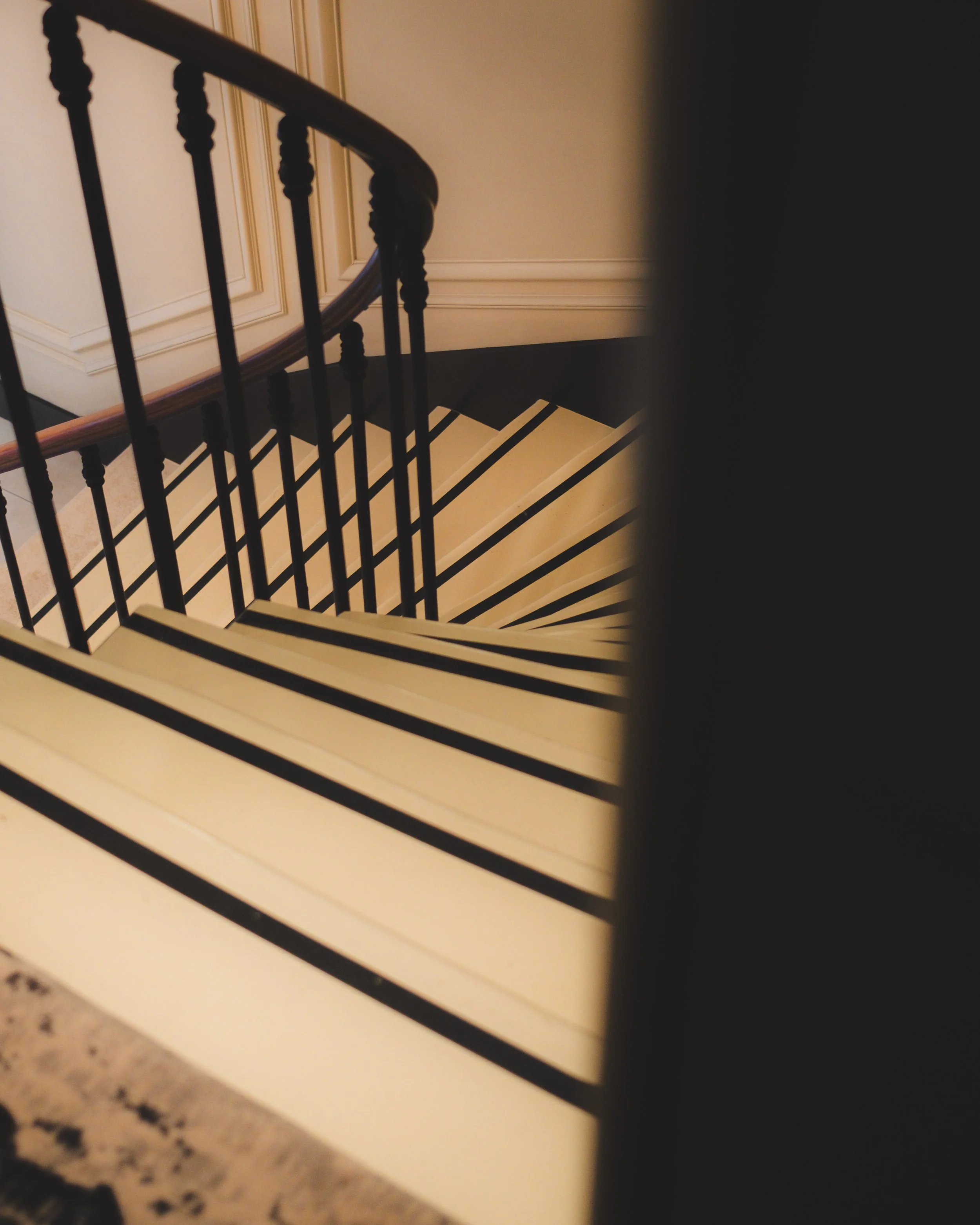 A spiral staircase ascending with black railings and white steps, viewed from above, with beige wall paneling visible in the background.