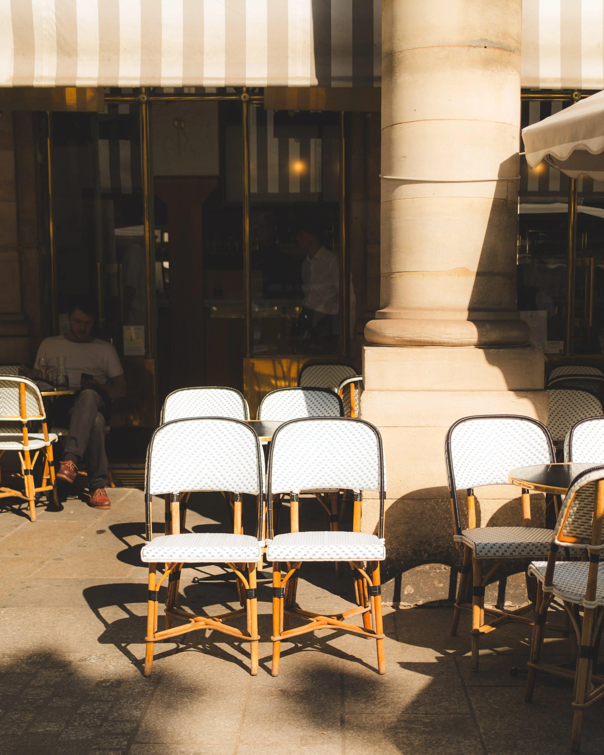 Outdoor cafe seating with several white woven chairs with black trim, yellow wooden legs, and intersecting supports. Substantial stone column and glass doors with brass framing in the background, with a person blurred inside and another person seated