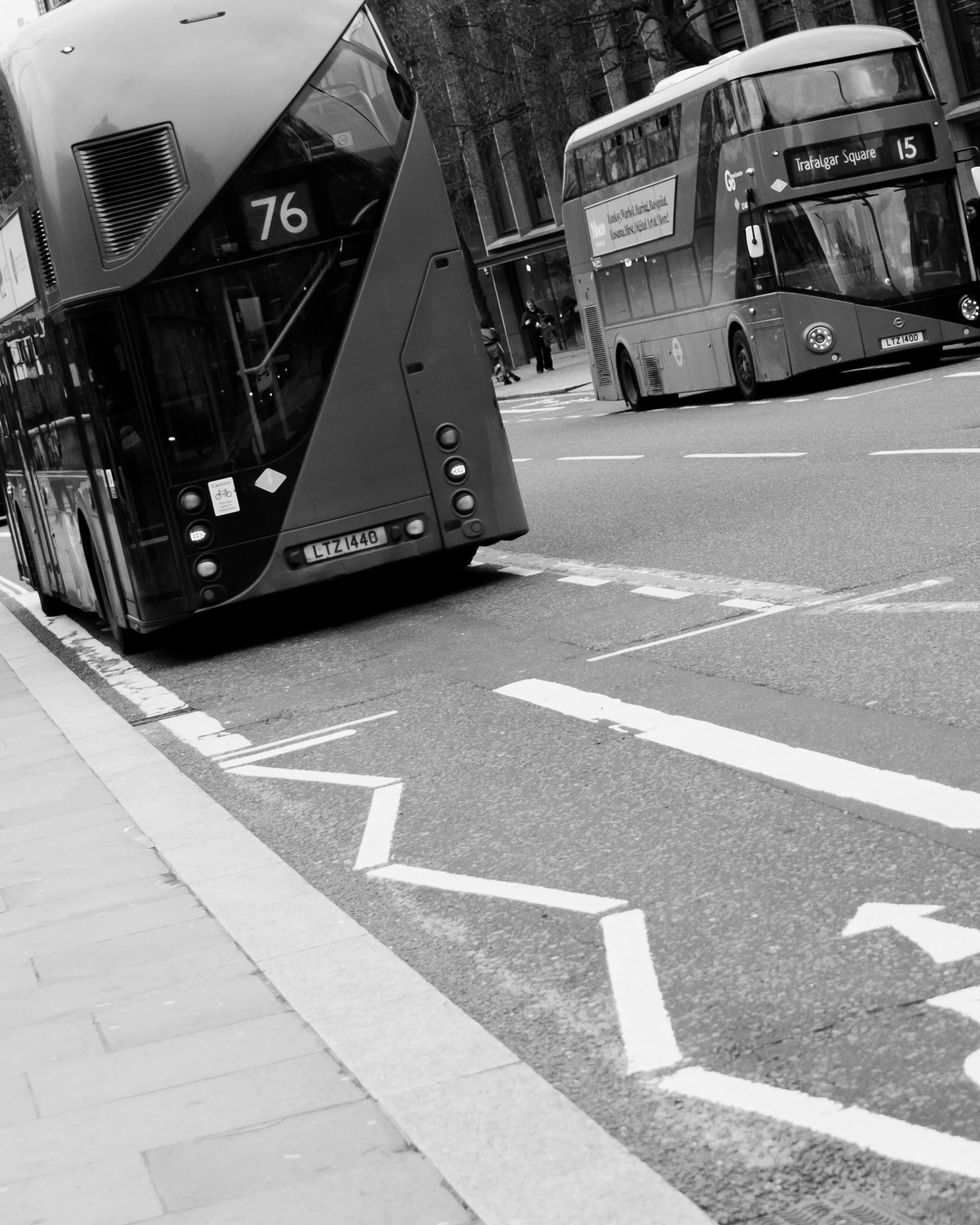 Black and white street scene with two double-decker buses, one parked and one in motion near a bus stop, alongside bicycle lane marked on the road, in front of a modern building. Lionel Chu - Hotel & Travel Photographer