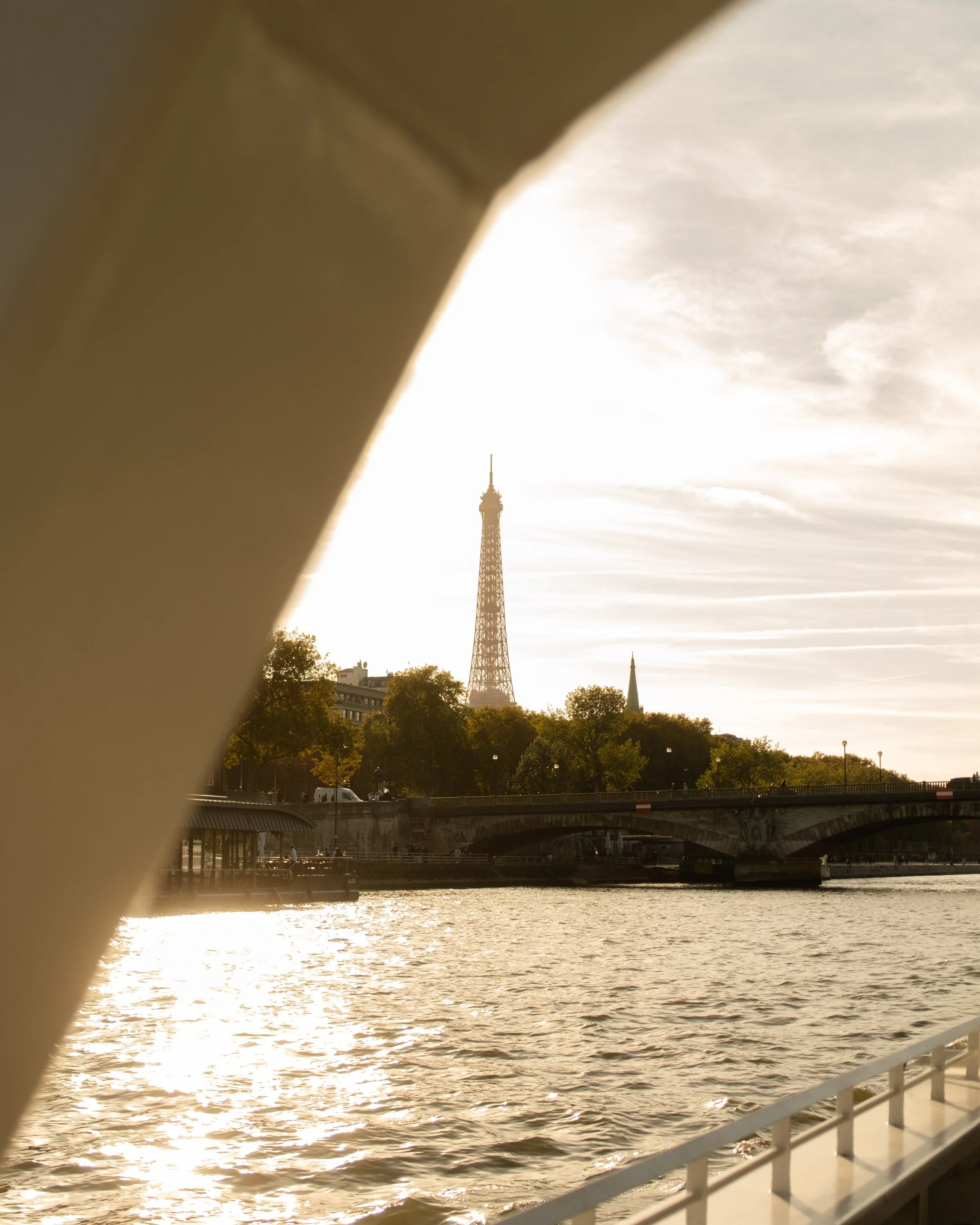 View of the Eiffel Tower through a boat window with water, trees, a bridge, and the Paris skyline in the background during sunset. Lionel Chu - Hotel & Travel Photographer
