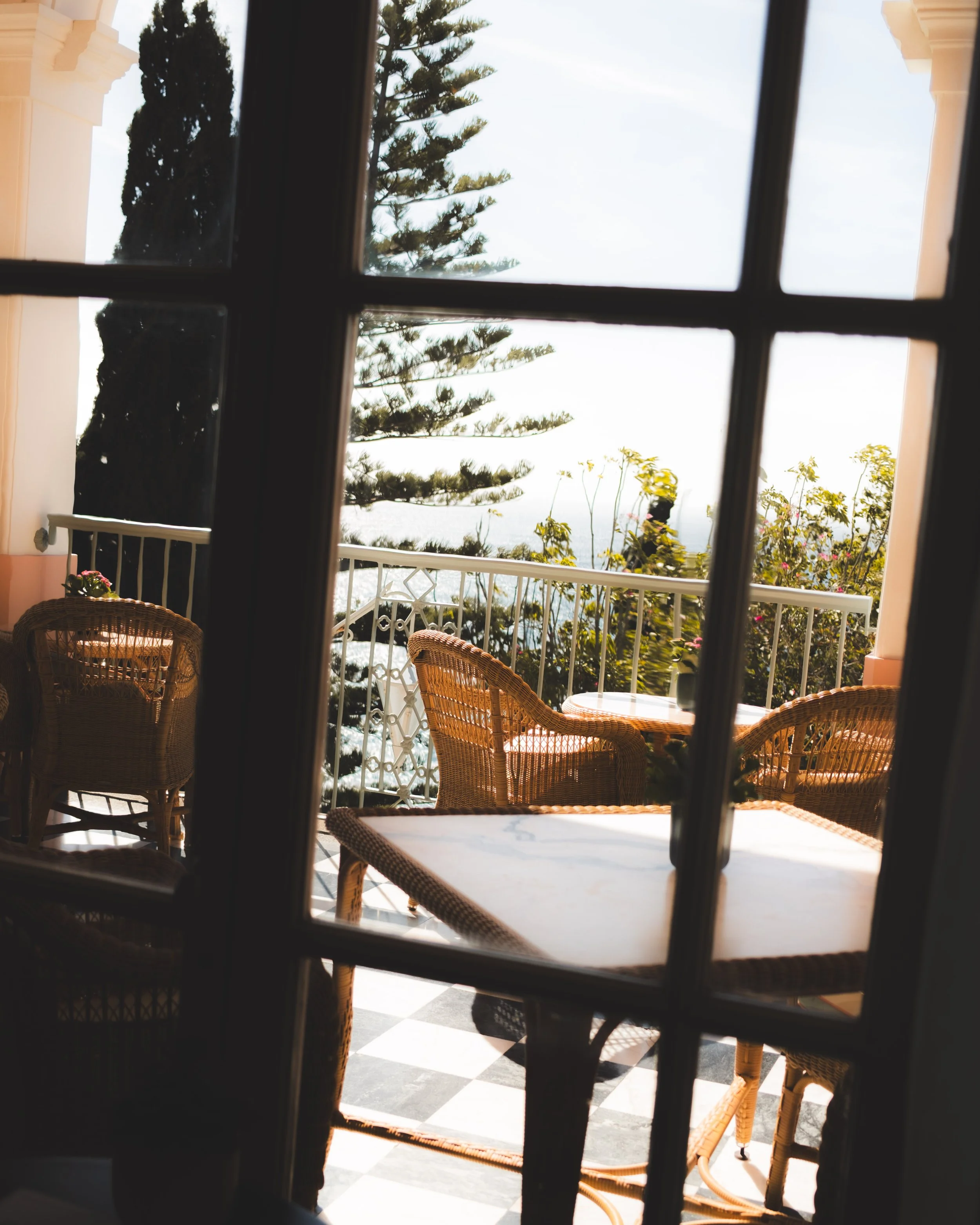 View through a window showing a balcony with wicker chairs and a table, a white railing, trees, and a cloudy sky.