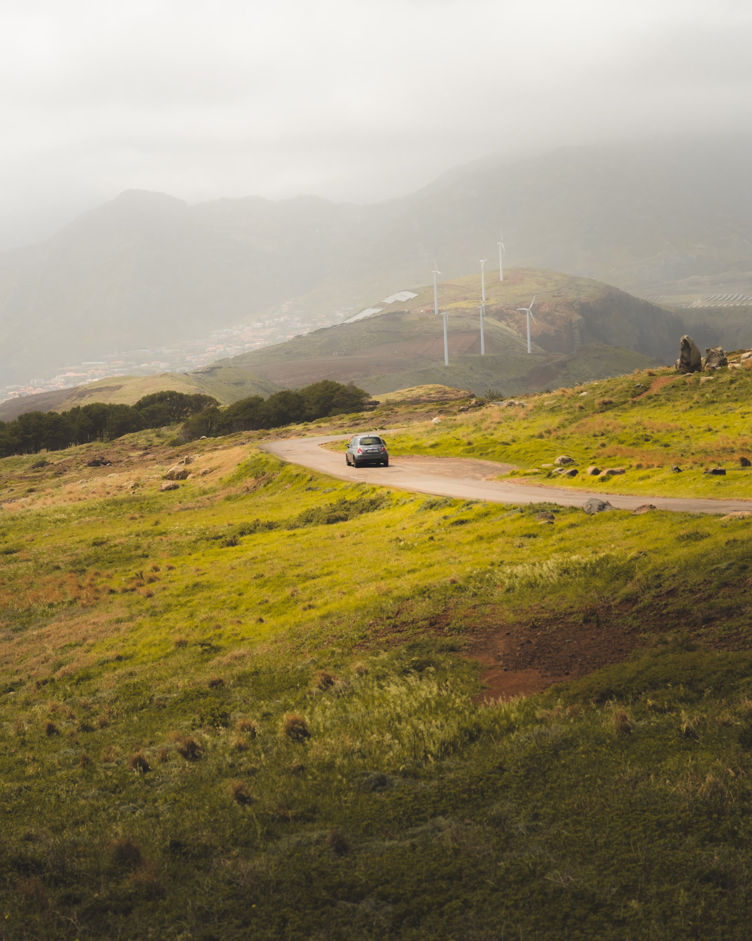A rural landscape with green hills, a winding dirt road, wind turbines on a hill, and a car driving along the road. Mountainous terrain in the background under cloudy skies.