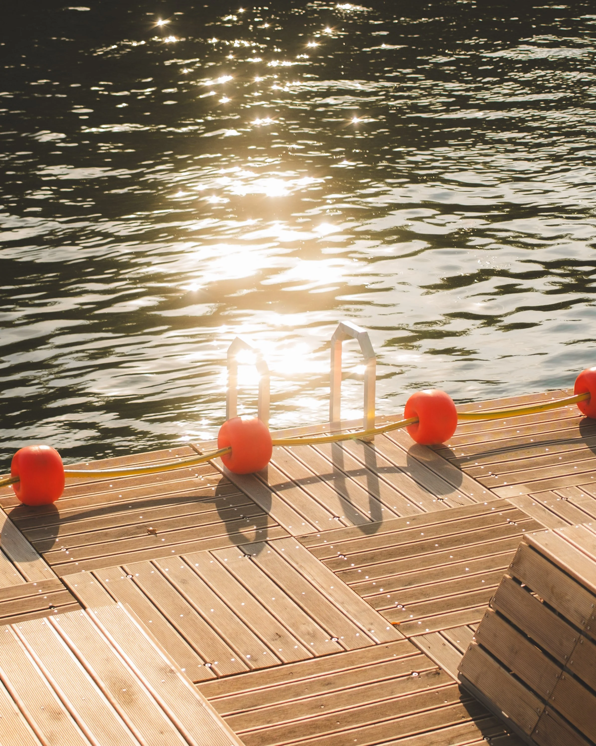 Sunlight reflecting off rippled water viewed from a dock with orange buoys and a floating ladder.