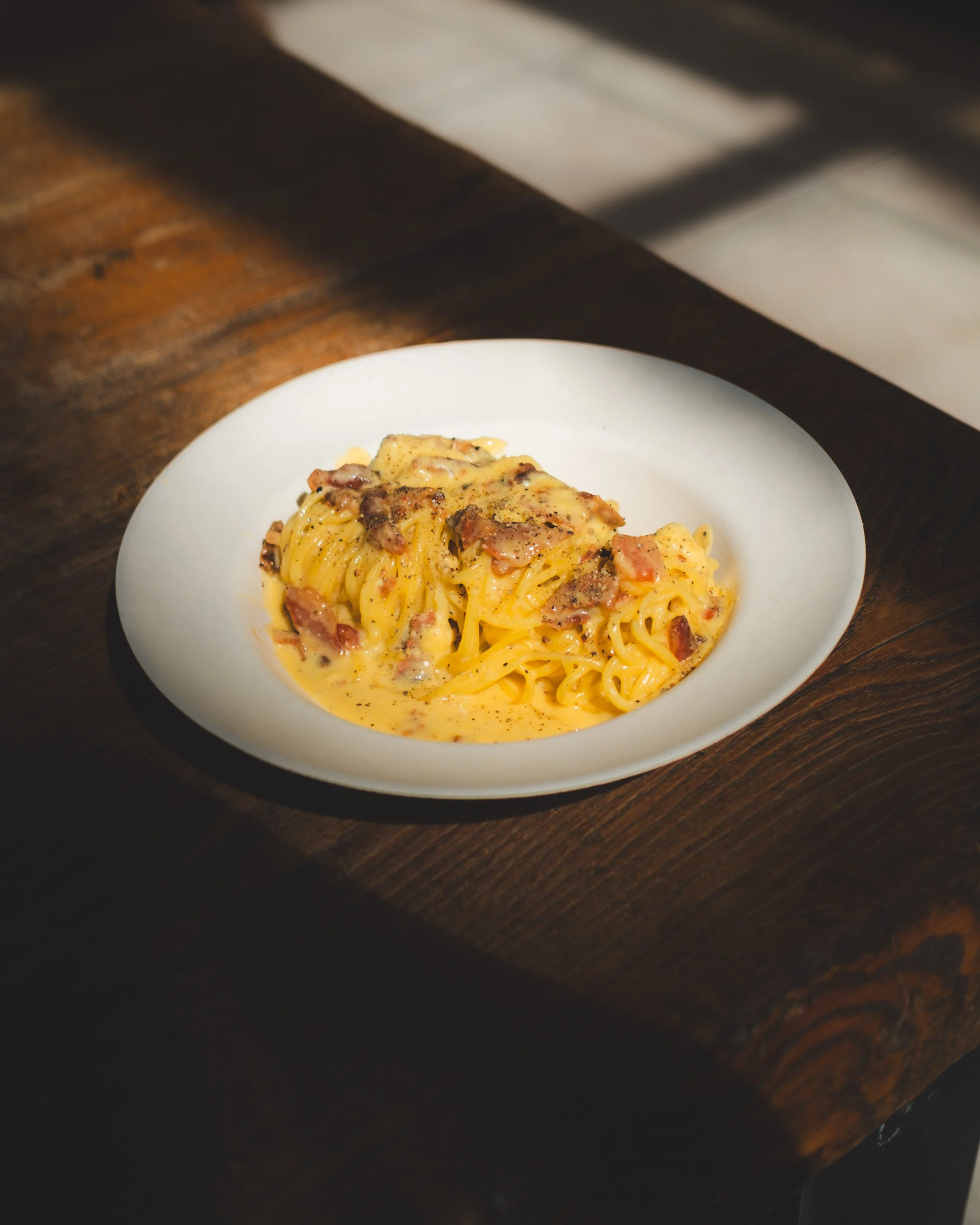 A white bowl of carbonara pasta with guanciale bits on a wooden table. Lionel Chu - Hotel & Travel Photographer