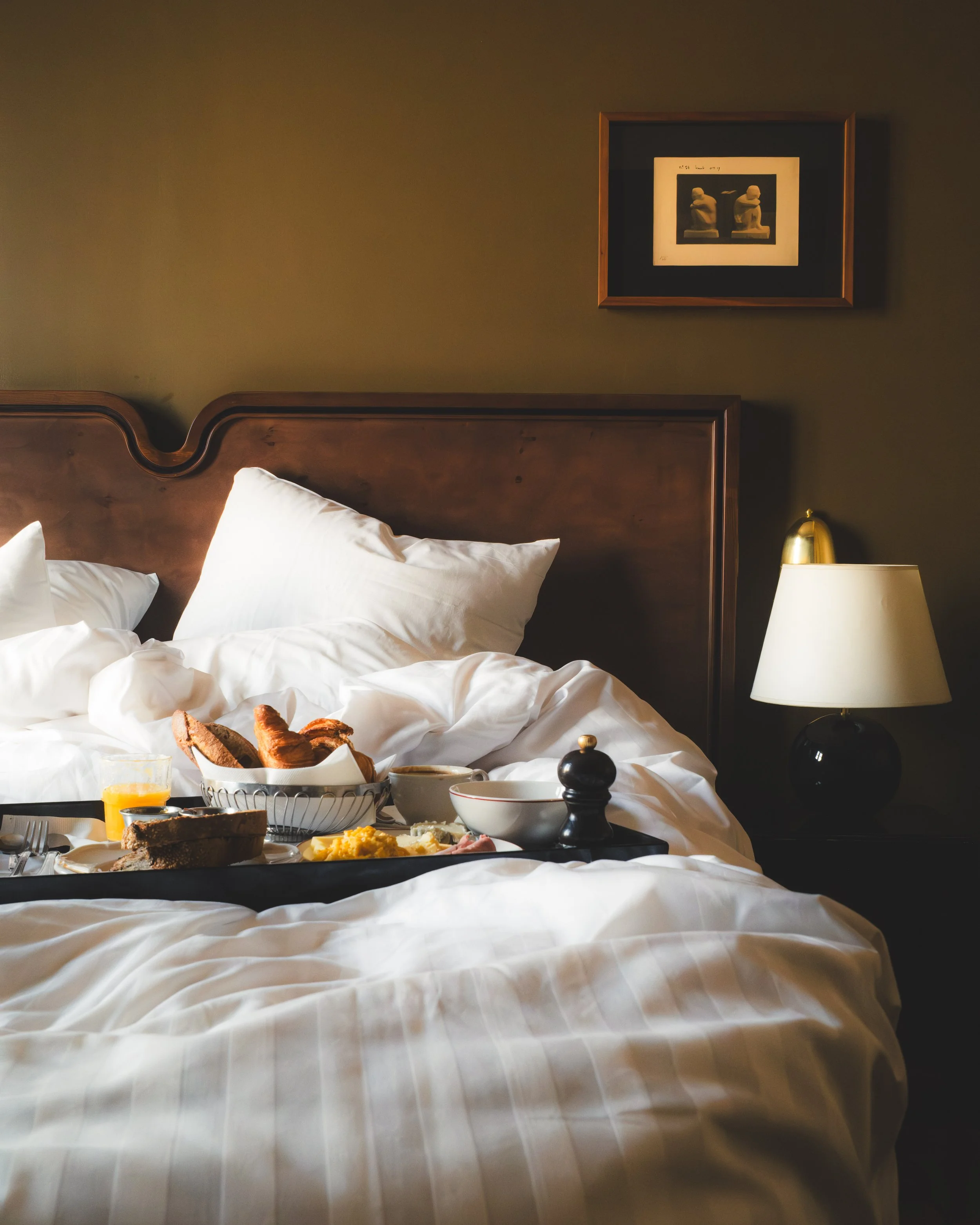 A breakfast tray on a bed with white bedding, featuring a bowl of scrambled eggs, slices of bread, and some breakfast meats, with a breakfast setup nearby including a glass of orange juice.