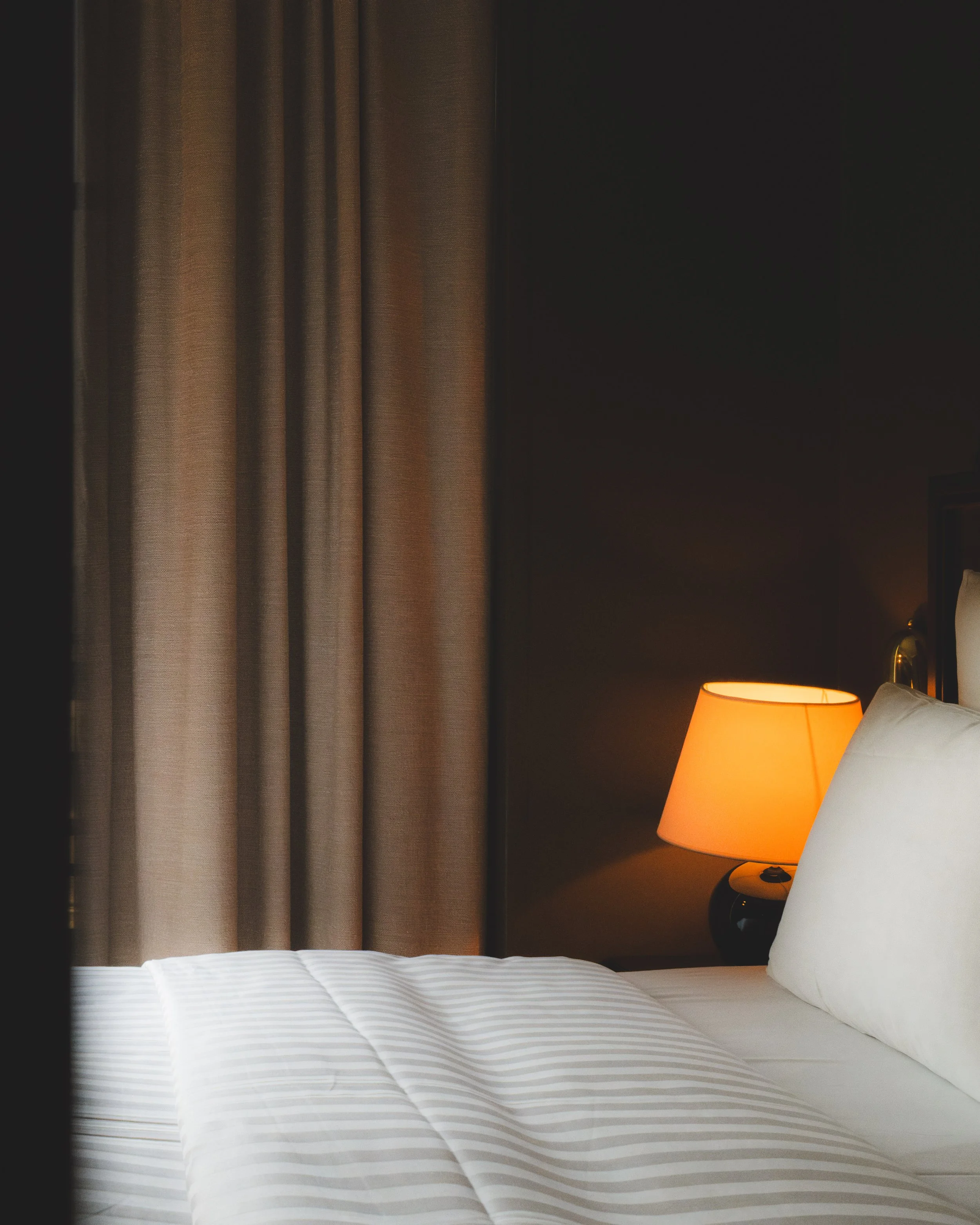 A neatly made bed with white striped sheets and pillows, beside a nightstand with an orange lampshade emitting warm light, in a hotel room with beige curtains.