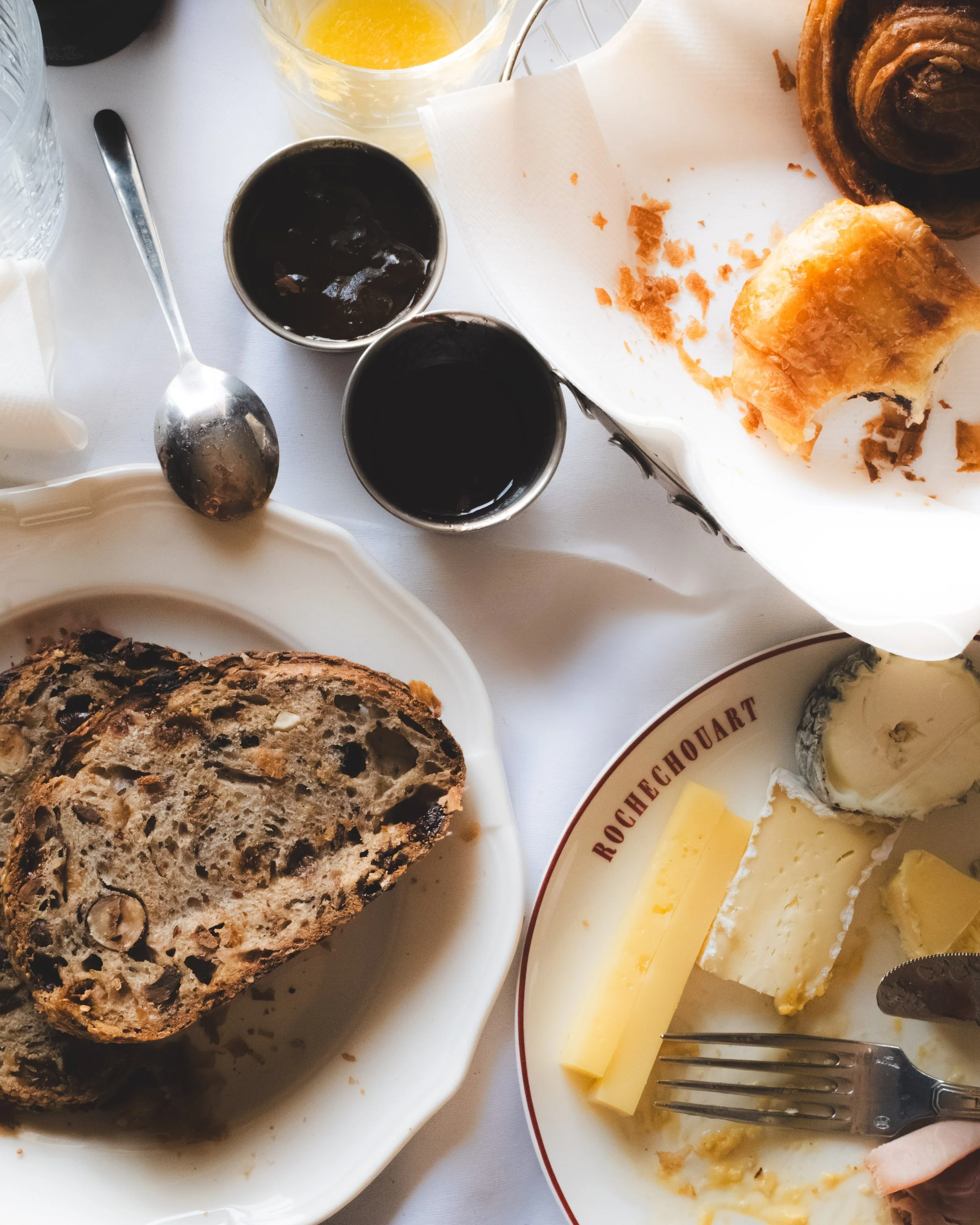 A breakfast table with slices of raisin bread, pats of butter, slices of cheese, two cups of dark coffee, a glass of orange juice, and a bowl of yogurt.