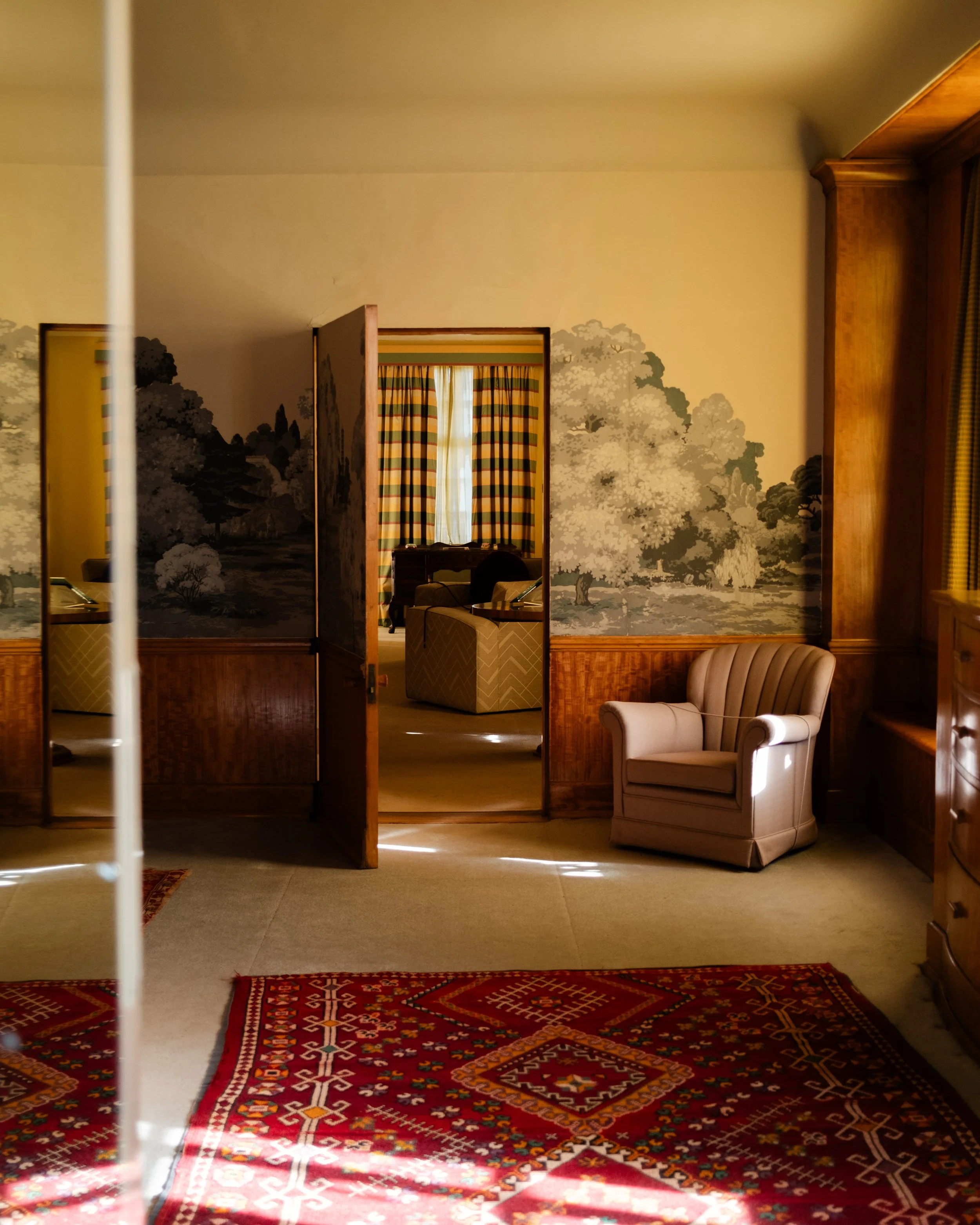An interior view of a vintage manor bedroom with a red patterned rug, beige armchair, and decorated wallpaper featuring trees and landscape scene, seen through an open door. Lionel Chu - Hotel & Travel Photographer