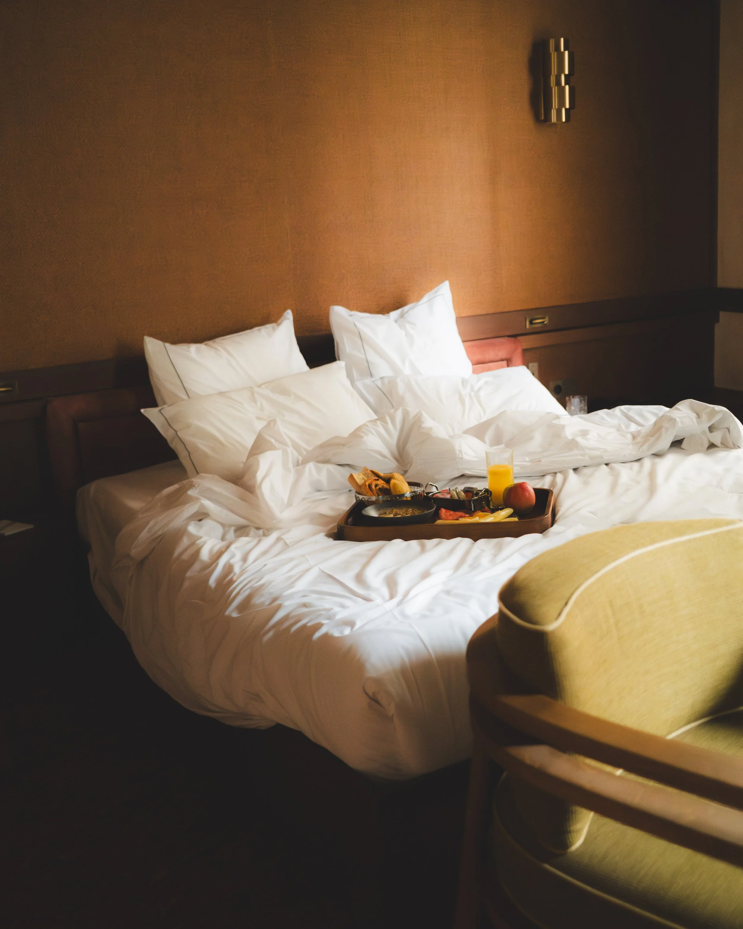 A hotel room with a bed, unmade with white sheets and pillows, a breakfast tray with food and drinks on the bed, and a beige upholstered armchair in the foreground.