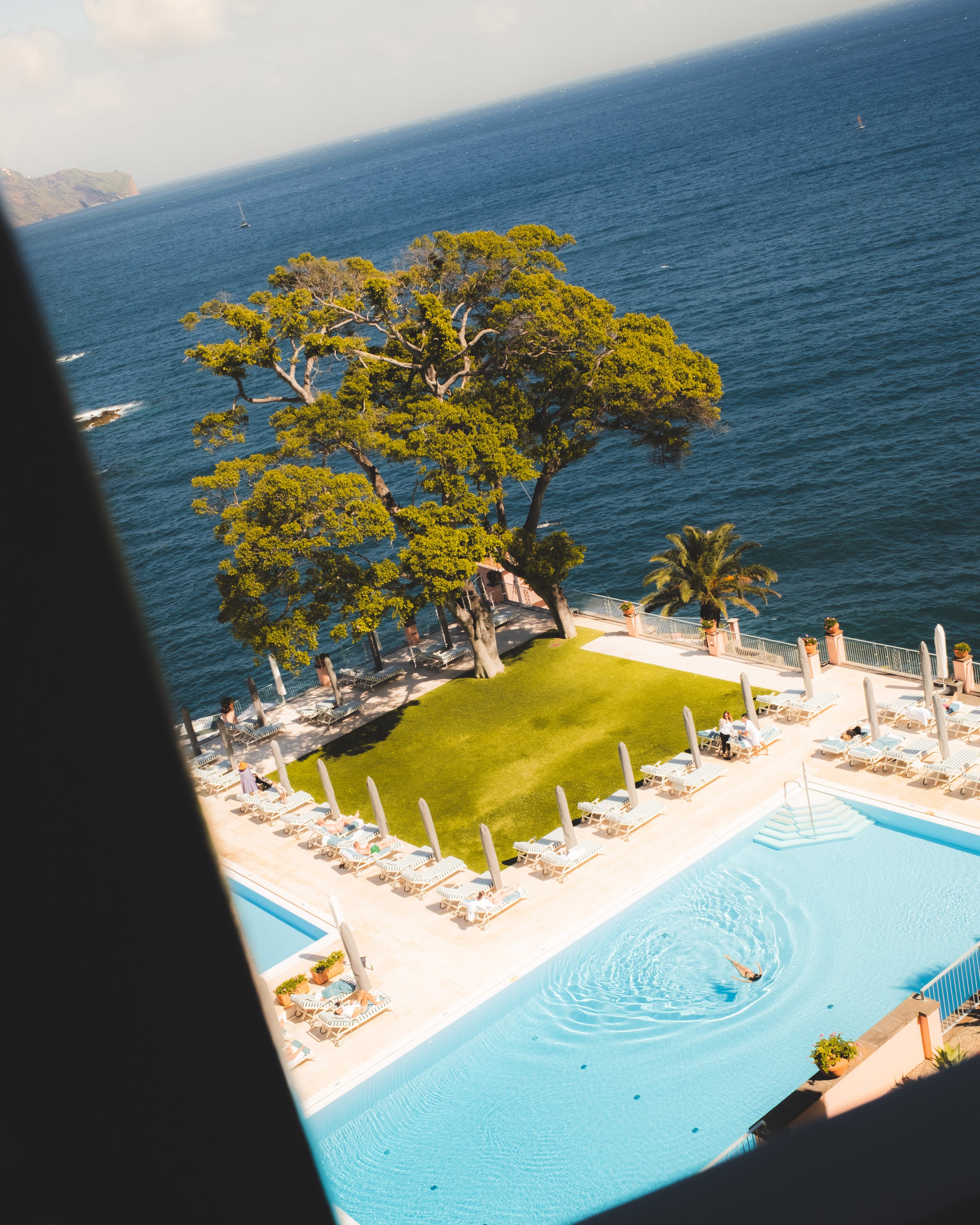 A view of a swimming pool with a person swimming, surrounded by lounge chairs, potted plants, and a fence, overlooking the ocean with boats and a coastline in the background.