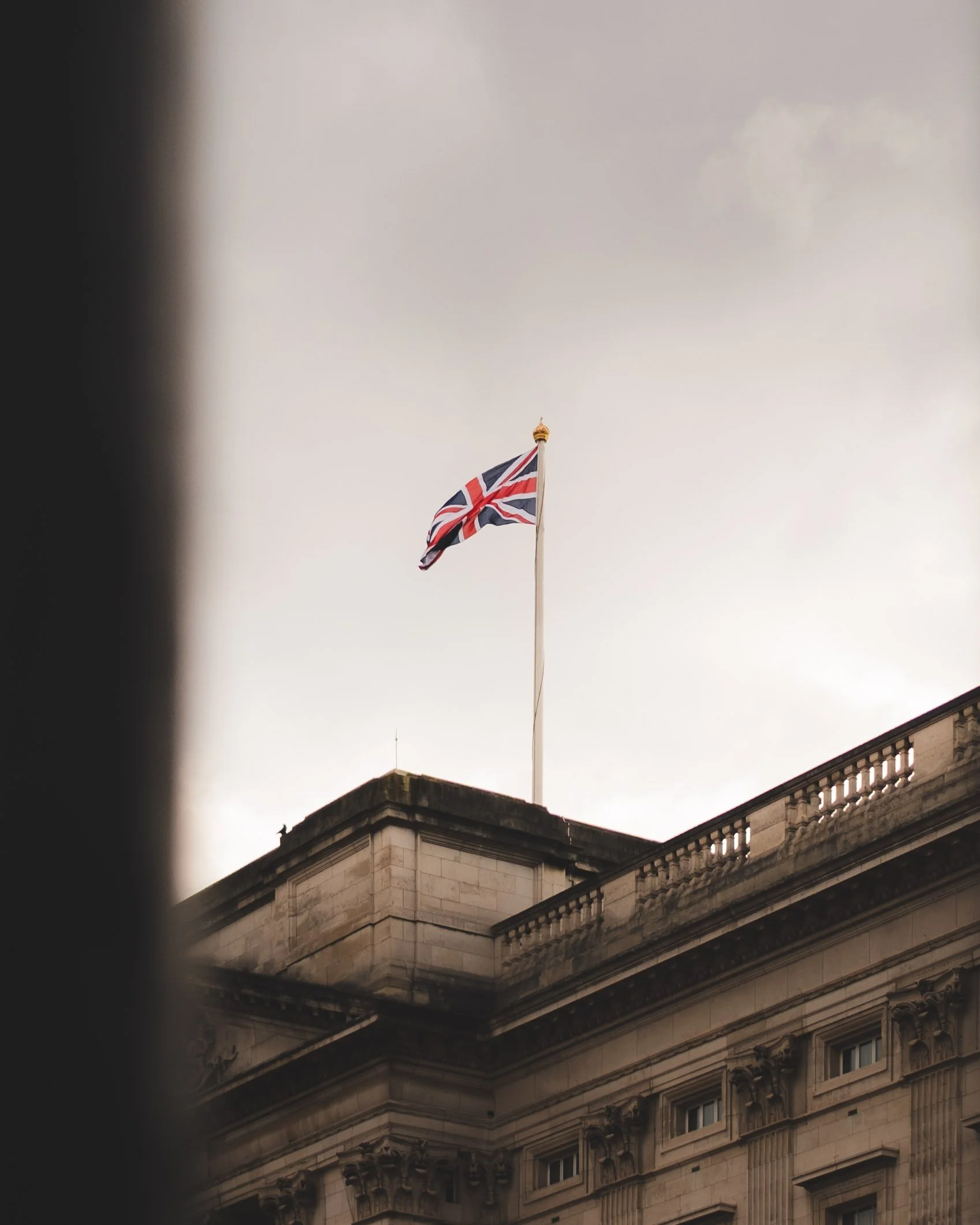 A flag of the United Kingdom flying on a flagpole atop a historic stone building with ornate architectural details, under an overcast sky.